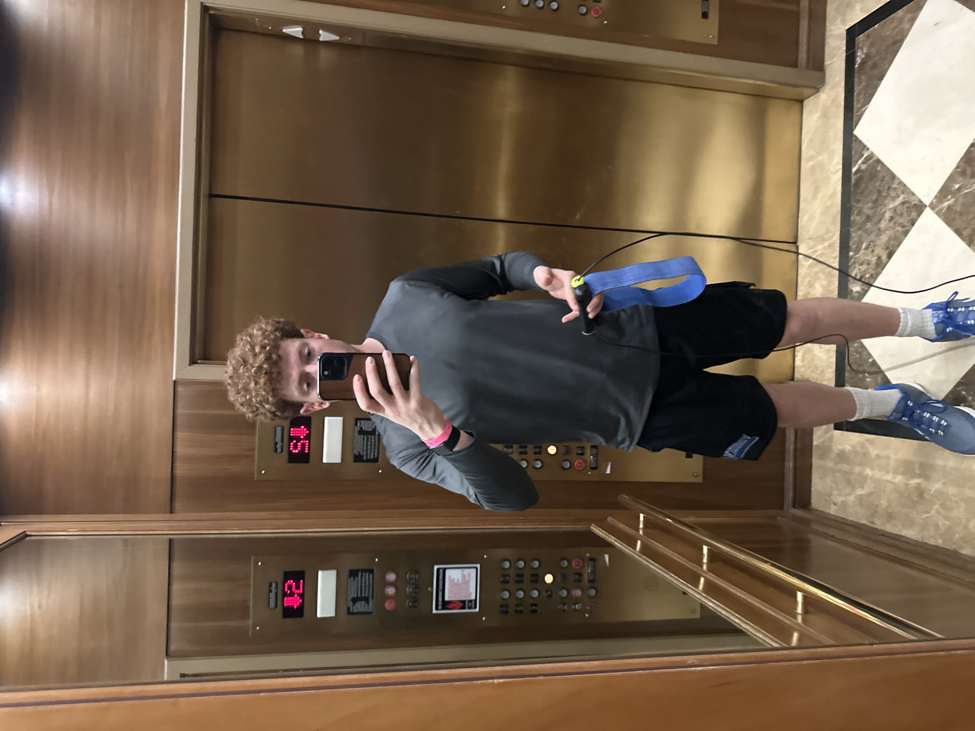 A person stands in an elevator, reflected in the mirrored walls. The elevator interior is predominantly gold with a control panel visible. The floor is a black and white checkered pattern. The person is holding a phone and a jump rope. The elevator is currently on the 12th floor, going up. It is unknown, without further information, if this is Aventura or any other city in the United States of America.