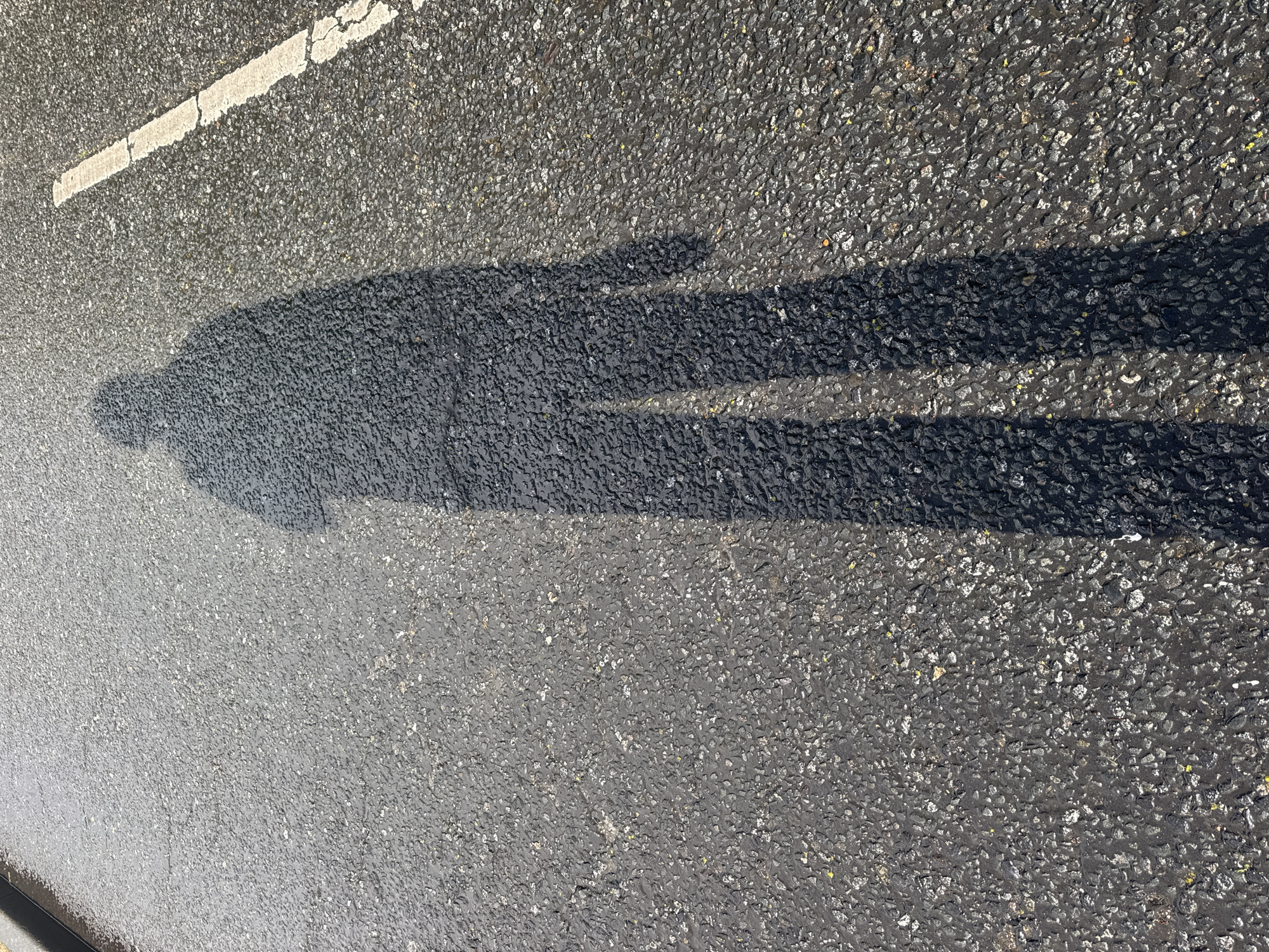 The image shows a shadow of a person on a wet asphalt road. A white line is visible on the left side of the image, indicating it may be a road marking. The location cannot be definitively determined to be London based only on the image content.