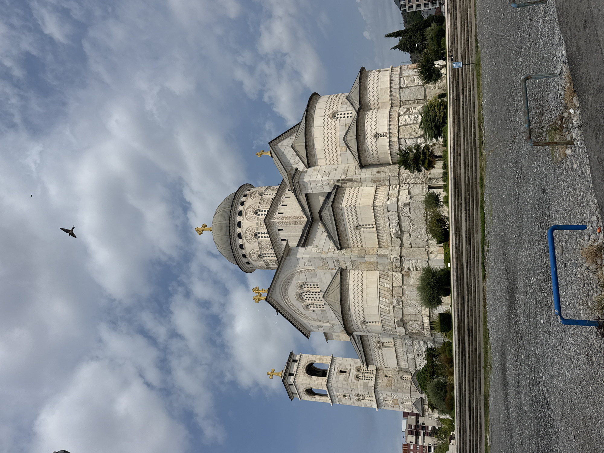 A white stone church with a large dome and bell tower is visible under a cloudy sky. A bird is flying in the sky above the church. In the foreground, there is a gravel area with a blue railing. There are stairs leading up to the church. The church has multiple domes and intricate carvings on its exterior.