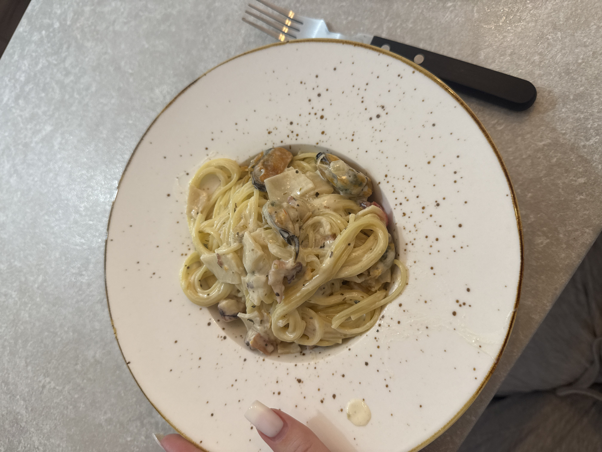 The image shows a plate of spaghetti with mussels and cream sauce, placed on a table. A fork with a black handle rests on the table beside the plate. A fingernail is visible in the lower left corner of the image. The plate is off-white with brown speckles and a brown rim. The background is a light grey patterned surface. The location is unspecified, but the prompt mentions Budva, Montenegro, although the image itself does not confirm this.