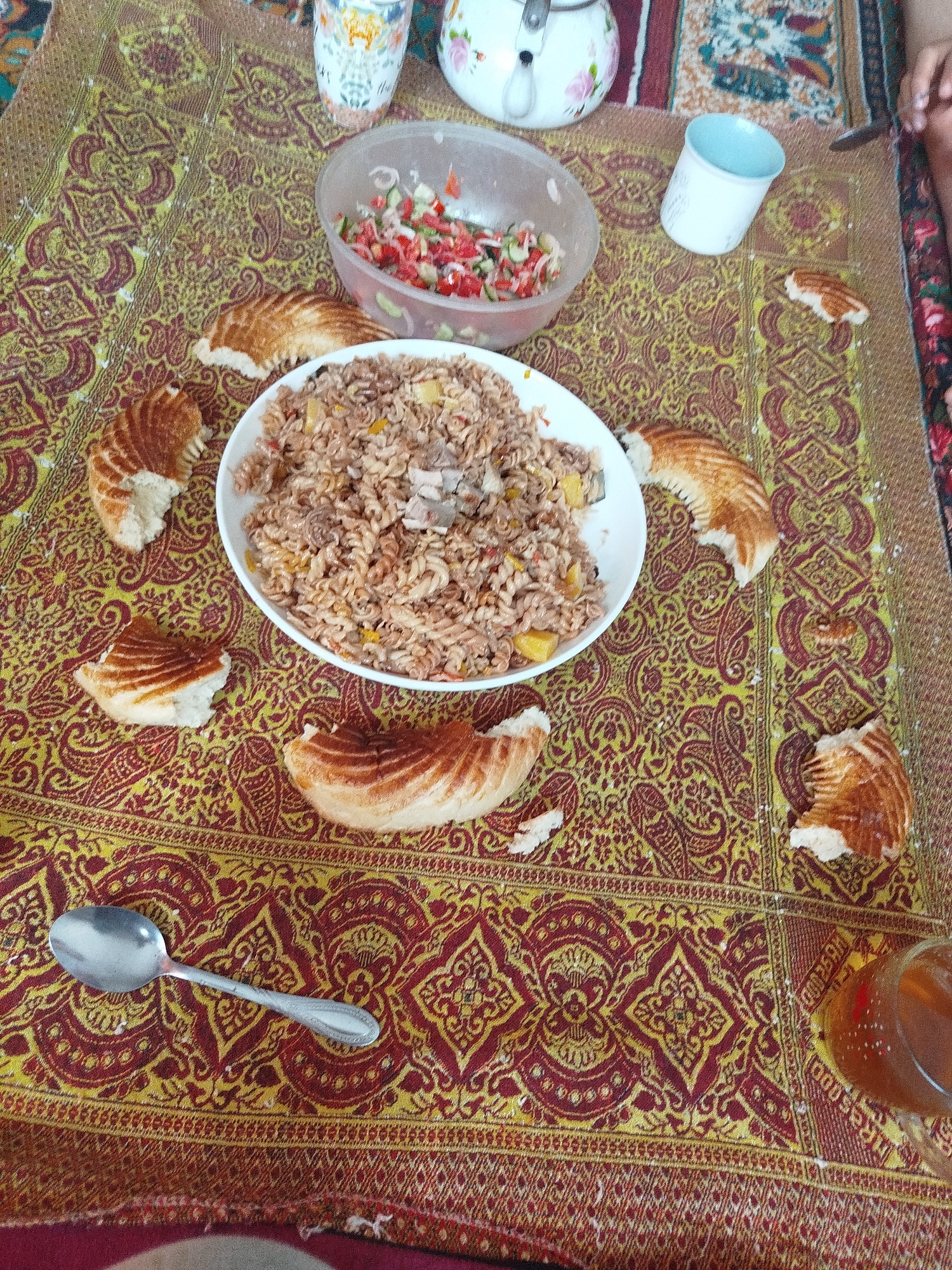 The image depicts a traditional meal laid out on a patterned textile, likely a tablecloth, in Aydarken, Kyrgyzstan. The meal includes a large bowl of pasta with meat and vegetables, a salad in a plastic bowl, and several pieces of flatbread arranged around the pasta. A silver spoon rests on the tablecloth near the bottom left of the frame. In the background, there are decorative cups and a teapot, suggesting a social gathering or mealtime. The overall setting appears to be indoors, possibly a dining area within a home, given the casual arrangement of the food and the presence of personal items.