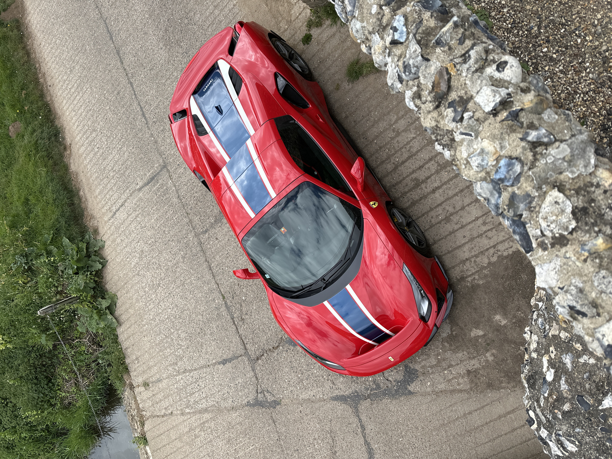 From an elevated perspective, a red Ferrari 488 Pista, embellished with a central racing stripe of blue and white, rests on a concrete surface. The car is parked near a stone wall and an area of vegetation. The location appears to be in a rural or suburban area within West Suffolk, United Kingdom. The view is slightly tilted and appears to be taken from a tall structure or a bridge. No people are visible.