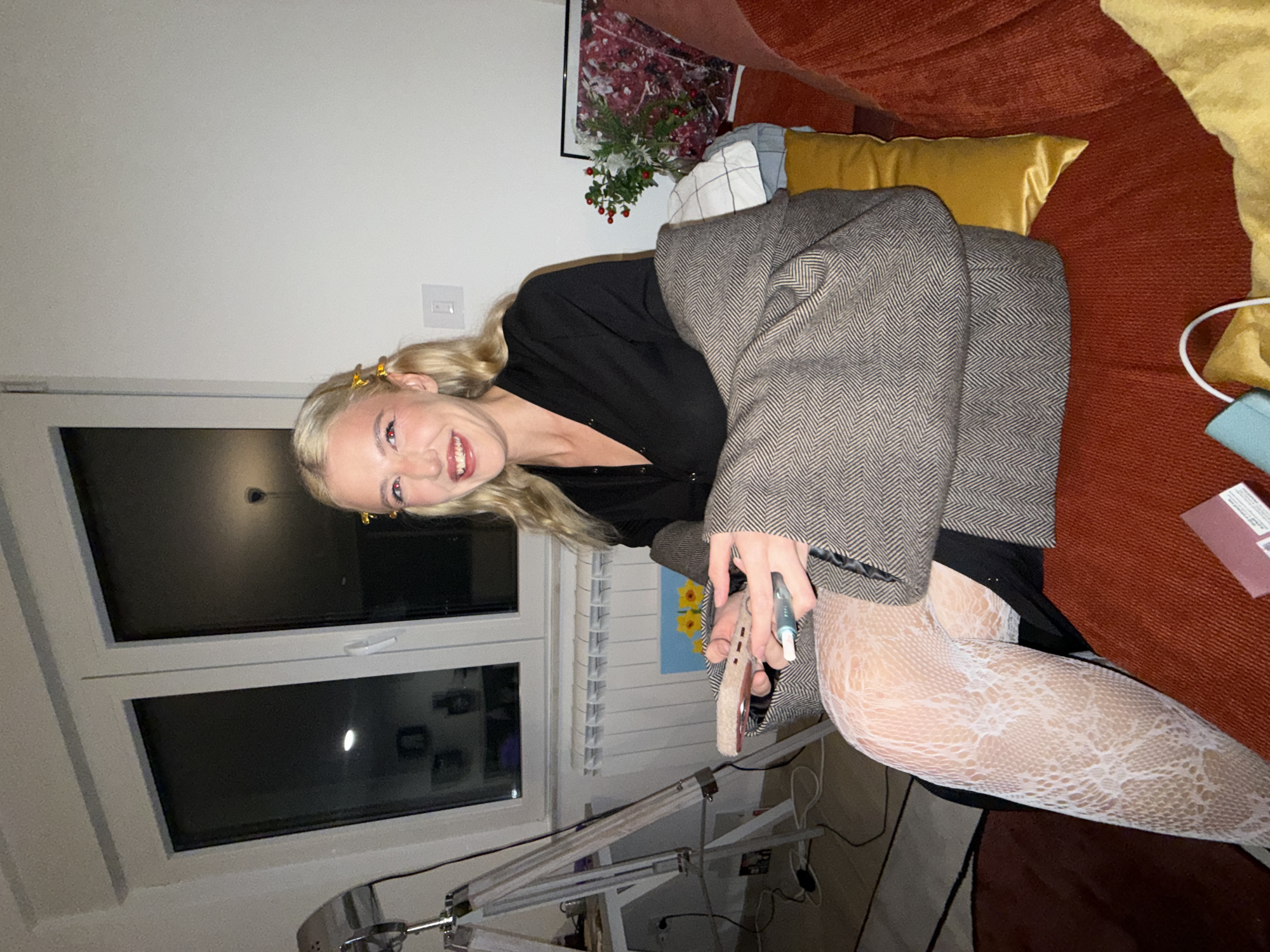 A woman is sitting on a red couch in a room with a window and a desk lamp. She is wearing white lace tights, a black shirt, and a grey herringbone blazer. She is holding a makeup sponge and a blue applicator in her hand. The location is not specified.