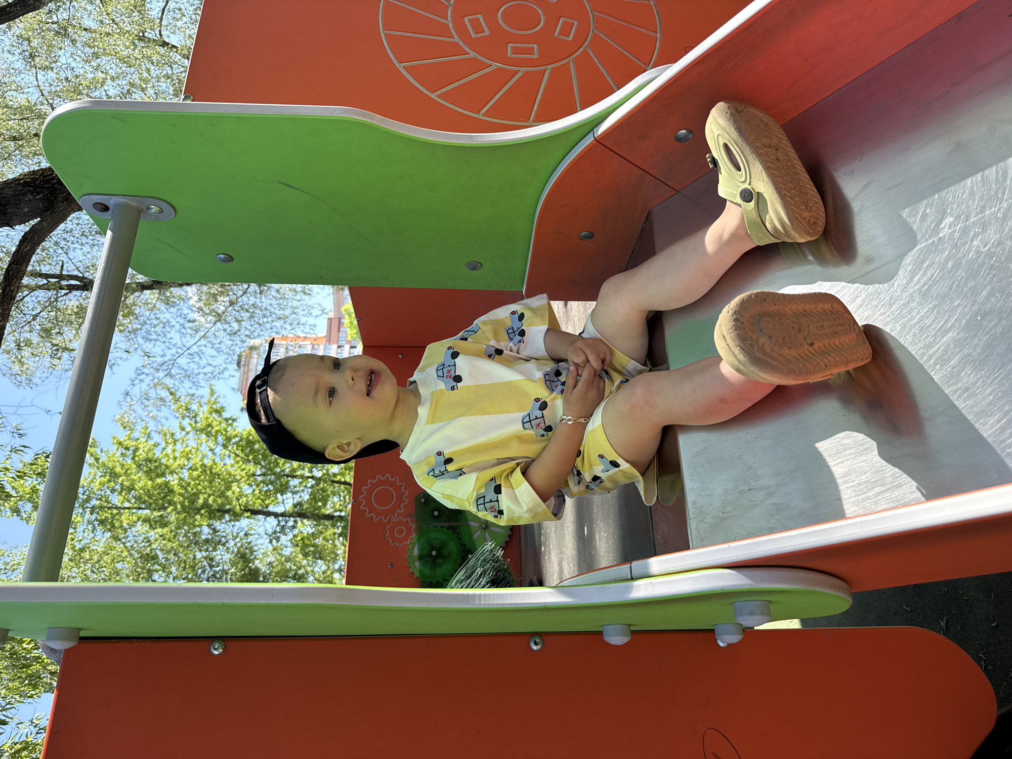 A young child, wearing a black baseball cap and a yellow and white striped shirt with blue car designs, is sitting on a silver slide at a playground. The child is wearing light-colored shoes. The slide is part of a play structure with orange and green elements. The playground is in an outdoor setting with trees and blue sky visible in the background. The location is Шебунино, Russia.