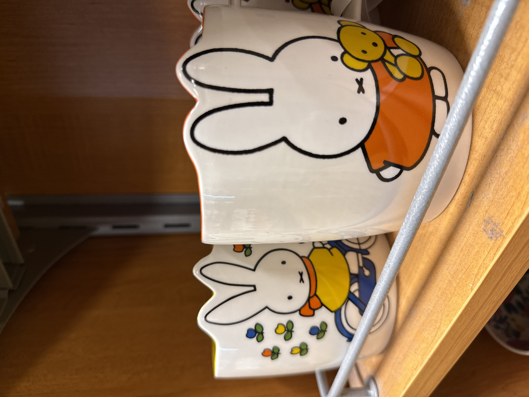 A close-up photograph taken indoors in Dania Beach, United States of America, shows two white mugs stacked one atop the other on a shelf. The mugs are decorated with an illustration of Miffy, a cartoon rabbit. The top mug depicts Miffy wearing an orange dress with yellow flowers, while the bottom mug shows Miffy wearing a yellow jacket and riding a blue bicycle, with small colored flowers scattered around. The mugs are separated by a wire rack. The shelf appears to be made of wood.