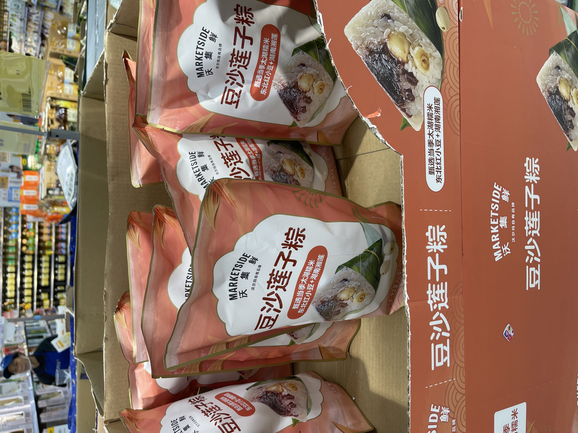 In a retail environment, likely a supermarket or grocery store in Panyu District, China, multiple packages of Marketside brand zongzi are displayed in cardboard boxes. The zongzi are a type of traditional Chinese rice dumpling, specifically featuring a filling of red bean paste, lotus seeds, and glutinous rice. Some packages are partially obscured by others. A person in a blue cap and apron is visible in the background, appearing to be a store employee. The lighting is artificial, typical of an indoor retail space. The foreground shows the consumer-facing packaging, with clear branding and product imagery.