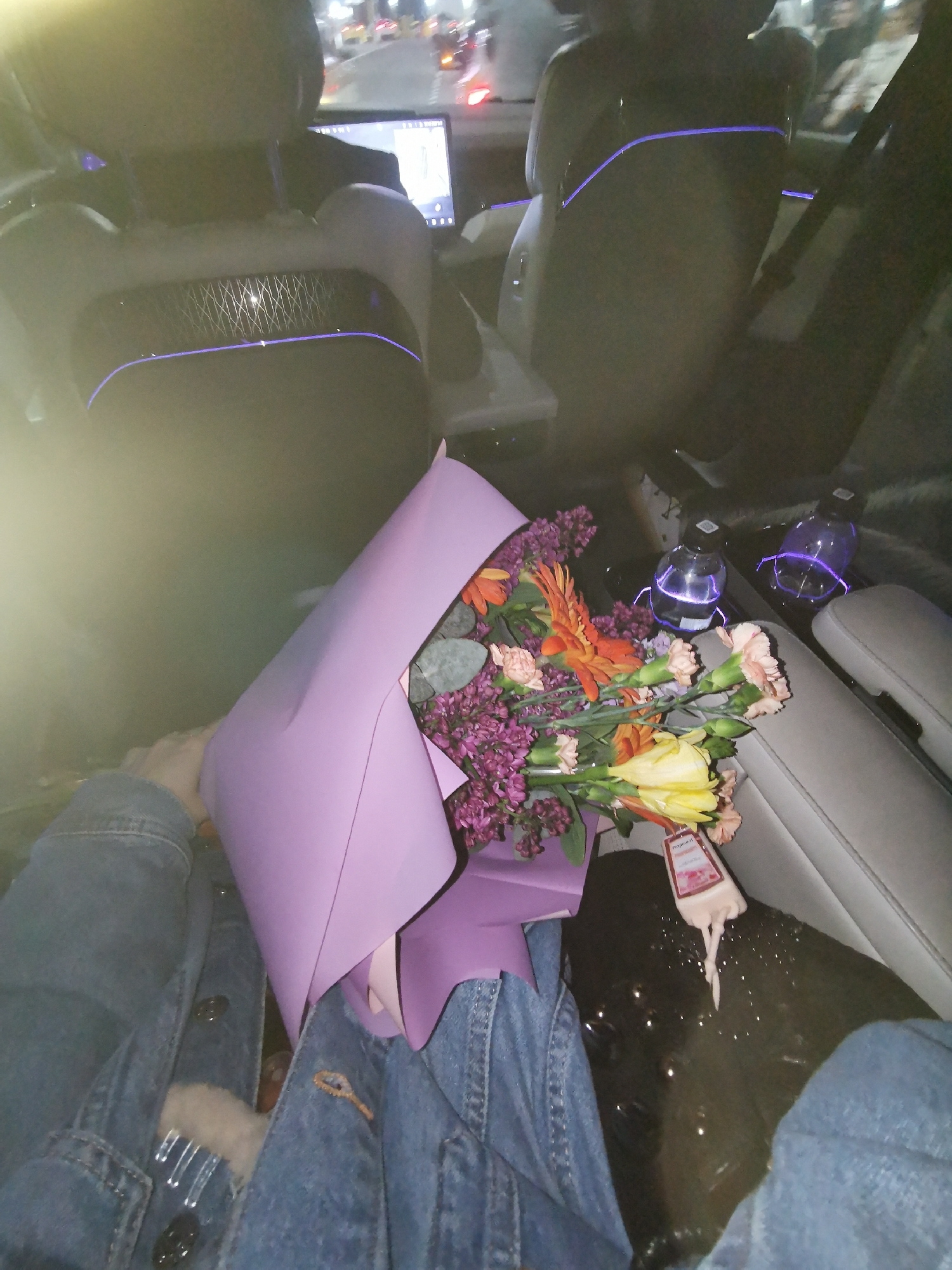 The image shows a person seated in the back of a car holding a bouquet of flowers wrapped in light purple paper. The flowers include orange gerbera daisies, purple lilacs, yellow tulips, and pink carnations. The person is wearing blue denim jeans with visible button closures. The car interior features illuminated drink holders with bottles, and a center console with a light-colored seat. The back of the front seats show a design pattern with blue accent lighting, and a screen is visible on the headrest of the front passenger seat. Out of the car window, streetlights and tail lights suggest a night-time setting within a city. The location is Unknown, Unknown.