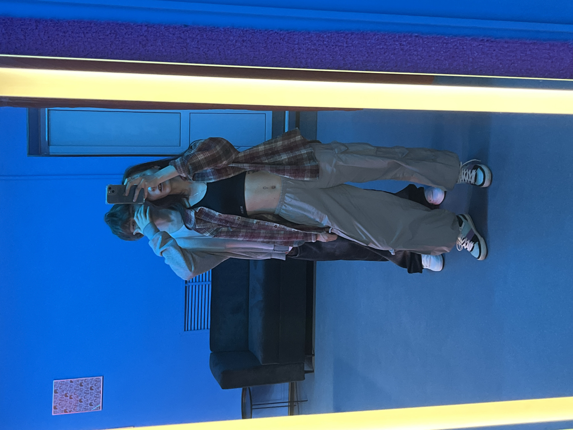 A vertically-oriented photo shows two people taking a selfie in a room painted in blue and purple hues. The primary individual, who is a woman, holds a smartphone to capture the image. She is wearing a black sports bra, tan sweatpants, and a brown and red plaid shirt. A small piercing is visible on her stomach. A second individual, possibly a man, is standing next to her and wearing a light gray hoodie and black pants. In the background there is a dark gray sofa, a window, a picture with geometric shapes, and a long rectangular light. The exact location within Krasnoyarsk, Russia is not discernible from the image.