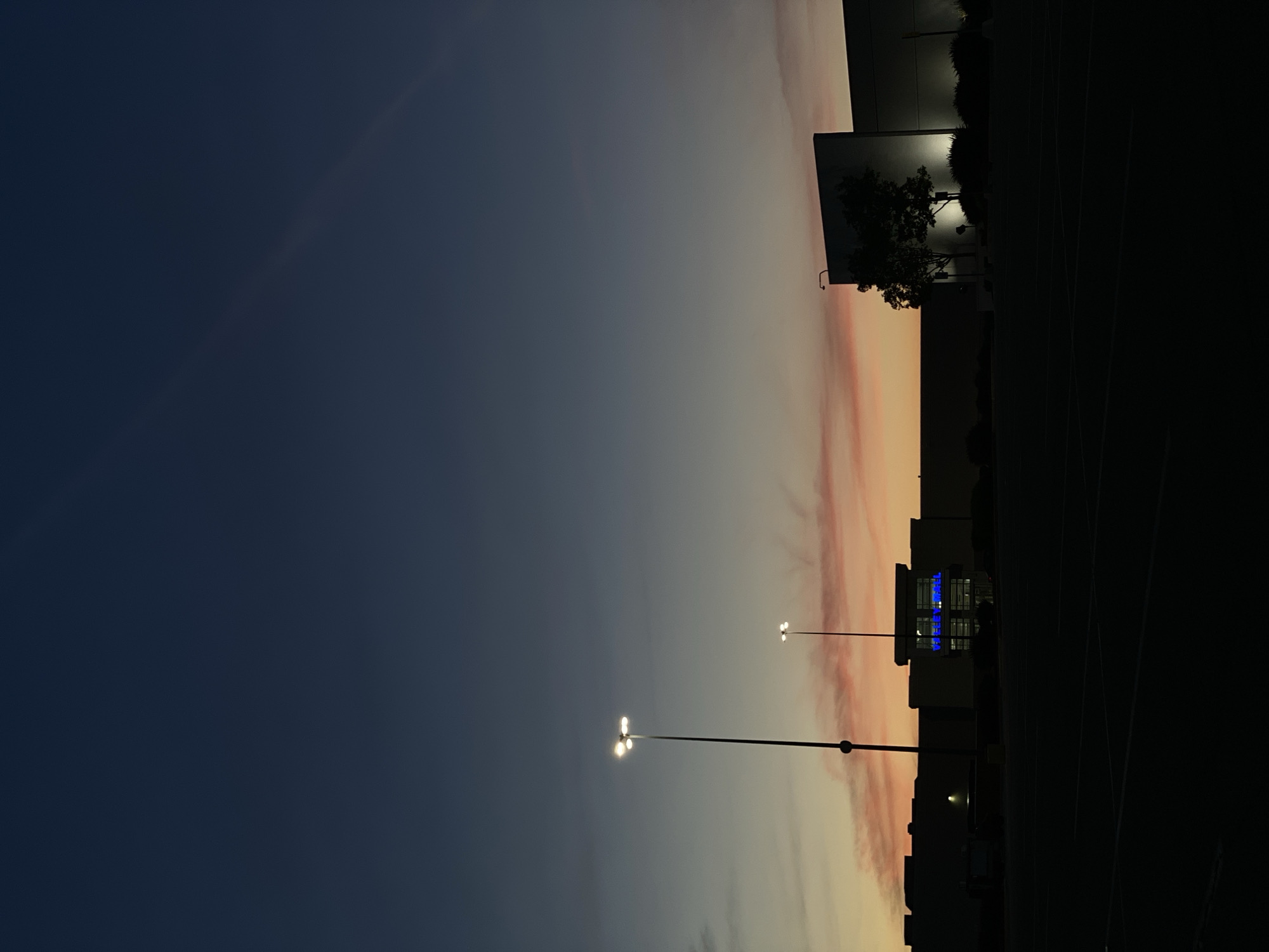 The image depicts a commercial building at dusk in Halfway, United States of America. The sky is a deep blue transitioning to lighter shades of orange and pink near the horizon. In the foreground, a dark, empty parking lot is visible. Two tall lampposts with multiple lights are illuminated. The building itself is silhouetted against the sky, with a section of its facade featuring bright blue illuminated signage that reads "VEETEN MALL". A single tree stands in front of another portion of the building, also silhouetted. The scene is devoid of people.