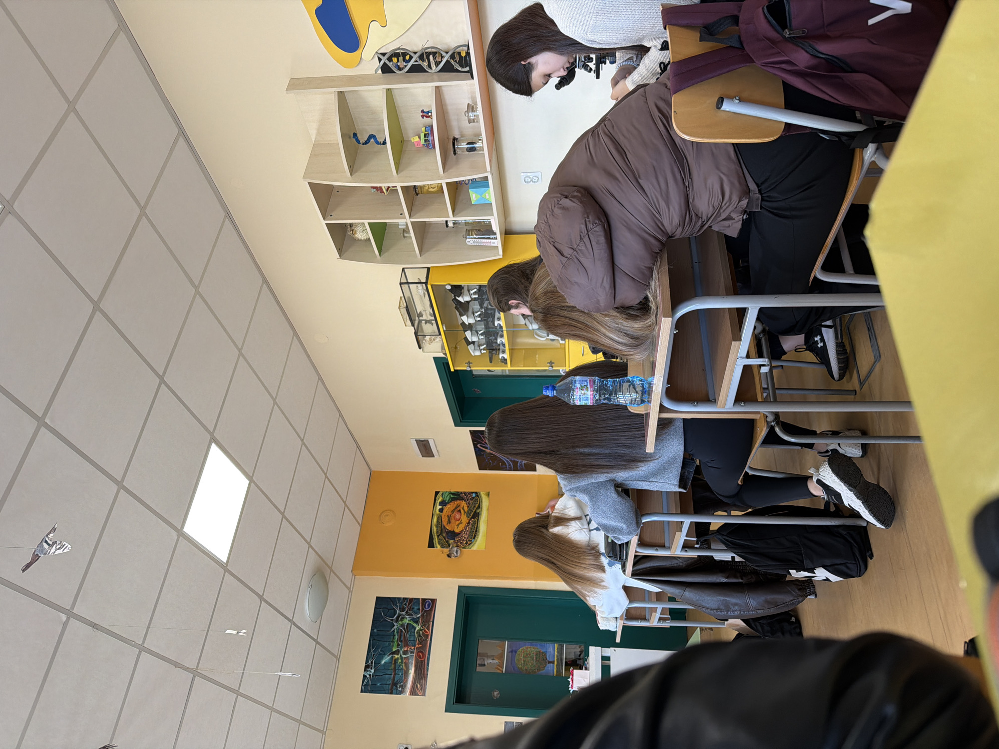 The image depicts the interior of a classroom in Panagyurishte, Bulgaria. Several students are seated at desks, appearing to be engaged in study or an activity. One student is looking through a microscope. Visible objects include desks, chairs, a bookcase with various items, wall art, a water bottle, and personal belongings such as jackets and backpacks. The classroom has a grid ceiling and pale yellow walls, with various pieces of decor throughout.