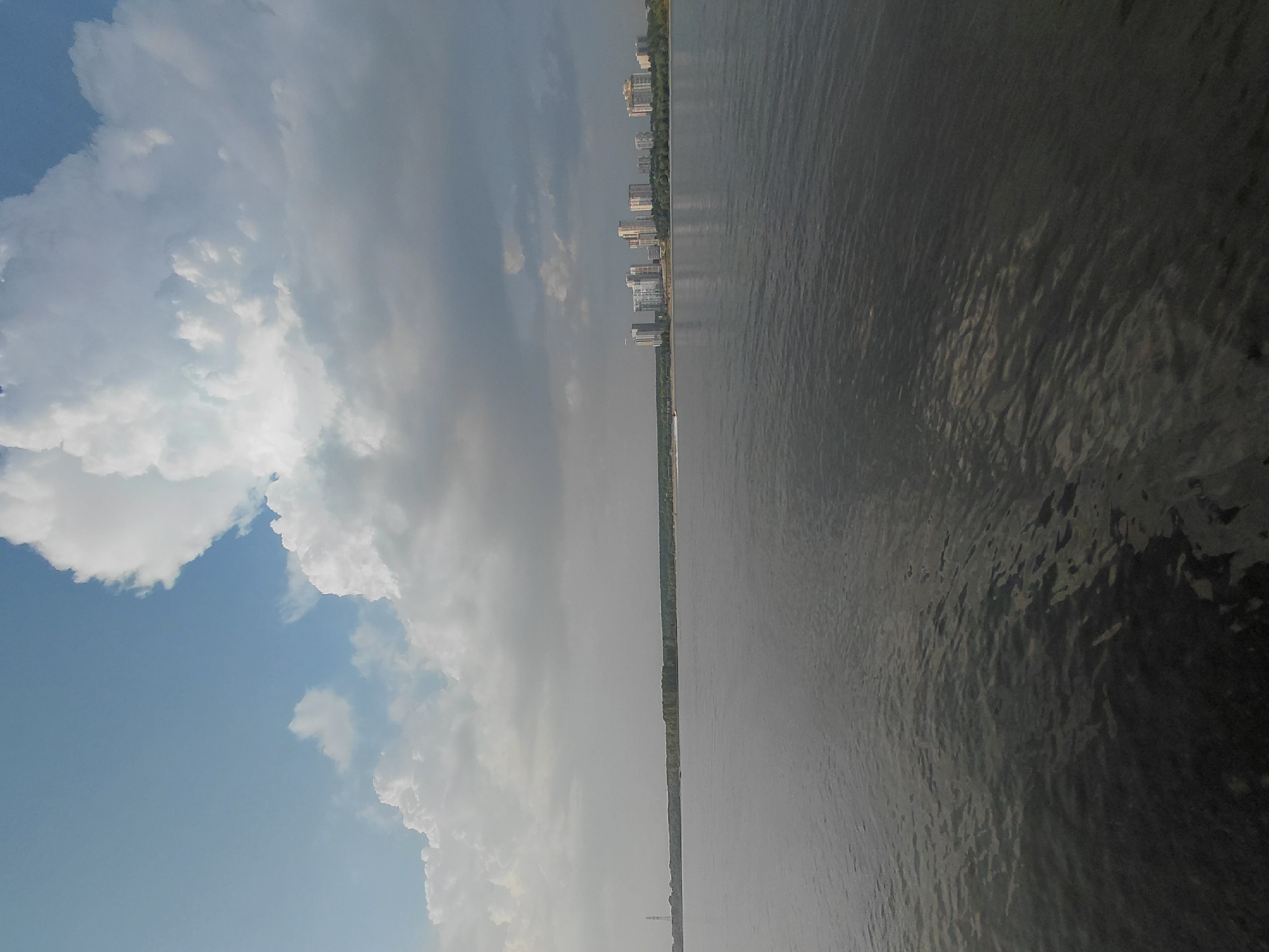 The image captures a wide river with a cityscape on the distant horizon, likely in Novosibirsk, Russia. The sky is dramatic, dominated by large, white cumulonimbus clouds tinged with grey, indicating a potentially changing weather pattern. Below the clouds, a hazy, light grey sky stretches across the scene. The river occupies the lower two-thirds of the frame, its surface dark and slightly rippled, reflecting the subdued light.
On the far bank of the river, a long, green treeline stretches across the landscape. Above the trees, a cluster of modern, multi-story buildings stands out, forming a distinct urban silhouette against the horizon. These buildings appear to be residential or office towers, characterized by light-colored facades. To the left of the main cluster of buildings, a tall, slender tower, possibly a communication or industrial structure, is visible. A small boat with a wake trailing behind it can be seen on the water in the middle distance. The overall impression is one of a vast natural expanse meeting an urban environment under a dynamic sky.