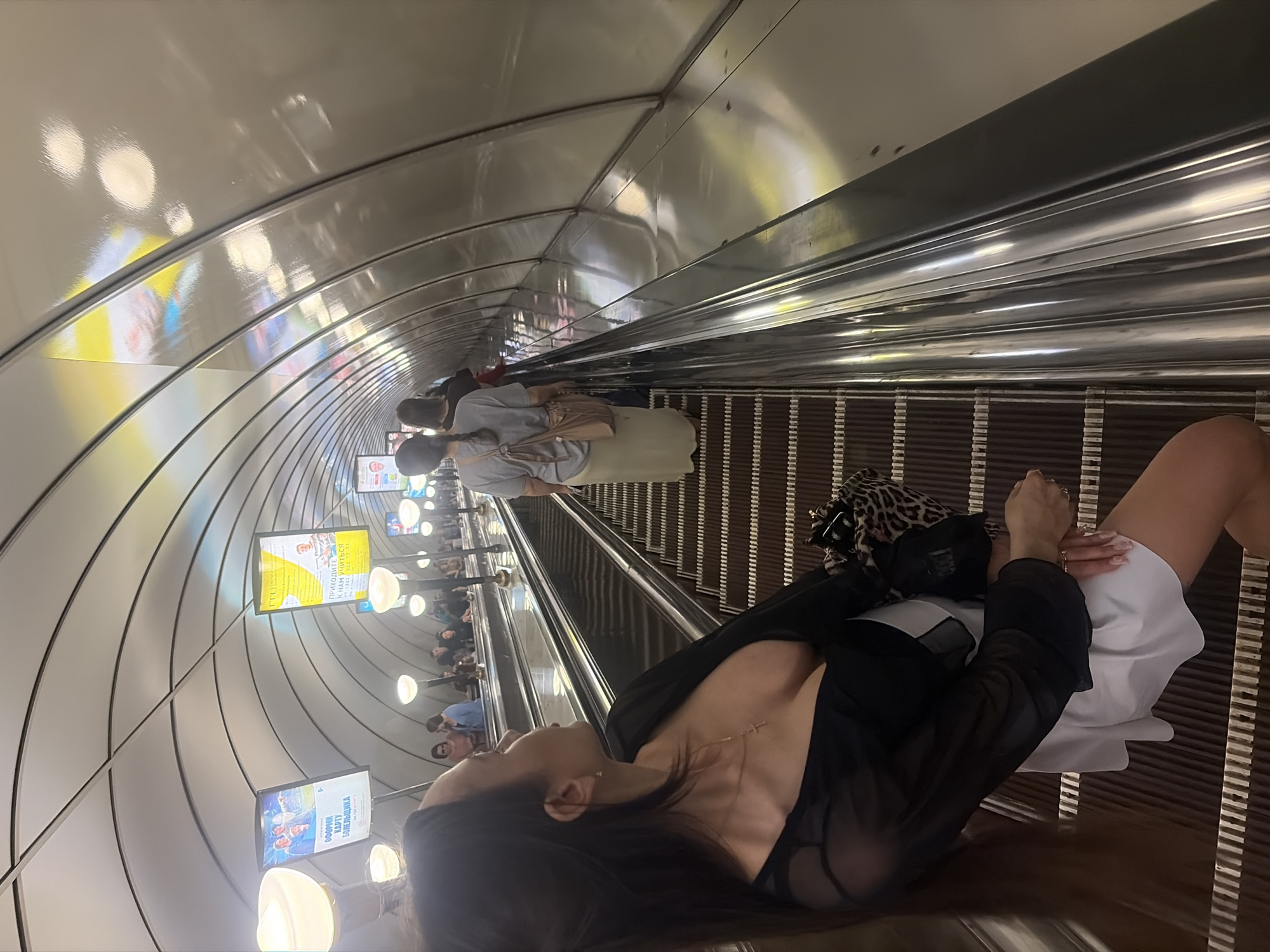 The image displays a deep escalator tunnel in the Saint Petersburg Metro, Russia. The tunnel walls and ceiling are constructed from light-colored, segmented, curved panels, forming a ribbed, arched structure. Illumination is provided by multiple spherical light fixtures mounted on poles between the escalators and by rectangular backlit advertising panels placed along the upper tunnel walls, displaying Cyrillic text such as "ОФОРМИ КАРТУ БОЛЕЛЬЩИКА" (Get a Fan ID) and "ПРИХОДИТЕ К НАМ УЧИТЬСЯ ГГЦ" (Come to study with us GGC).
Approximately ten individuals are visible, primarily ascending the escalators. In the foreground, on the right escalator, a woman with long, dark brown hair tied in a ponytail is shown in profile, looking upwards and to her left. She is wearing a black sheer long-sleeved top with a deep V-neckline over a white garment, and a short white skirt, revealing bare legs. A thin gold chain with a small cross pendant is visible around her neck. Her left hand, featuring manicured nails, rests on a small, leopard-print handbag. Behind her, a woman with dark hair in a braid is seen from the back, wearing a light grey t-shirt, a long light beige skirt, and carrying a light brown cross-body bag. Other commuters, indistinct due to distance, are further up the escalator. The escalators feature dark brown corrugated steps and reflective metallic handrails. The scene is brightly lit.