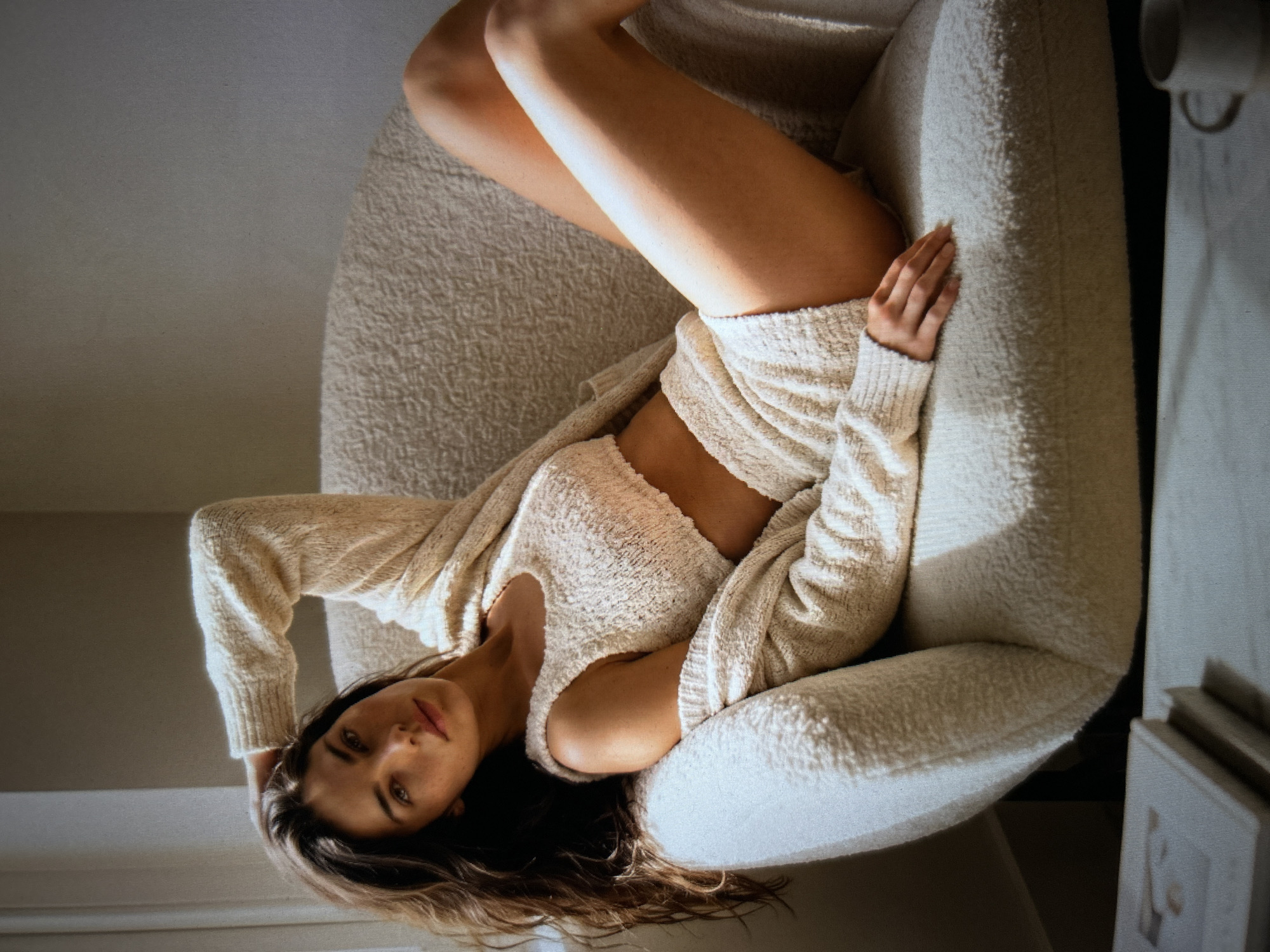 In an indoor setting with natural light streaming from the left, a young woman with long, dark hair is reclining on a textured, cream-colored armchair. She is wearing a matching cream-colored knit outfit consisting of a cropped tank top and shorts, with a cardigan draped over her arm and shoulders. Her right arm is bent behind her head, and her left hand rests on her lap. Her legs are crossed, with her right leg extending across her left. The background consists of a neutral-toned wall. In the lower-left corner of the frame, a stack of books is visible on a surface, and in the lower-right corner, a white mug is present. The lighting creates shadows and highlights on the subject and her surroundings.