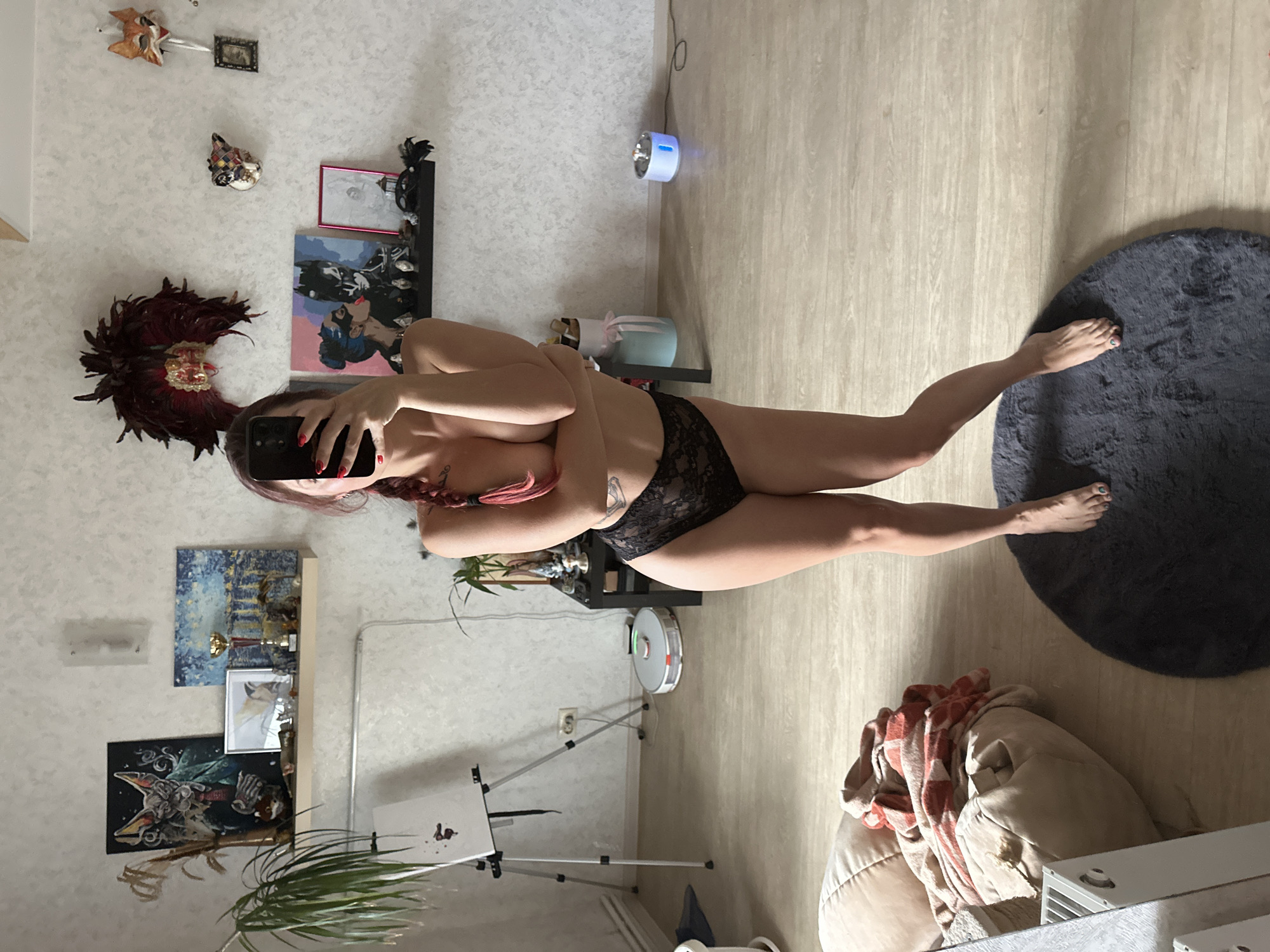 This is a vertically oriented, full-body mirror selfie of a woman in Vladimir, Russia. The woman is wearing black lace lingerie and has red hair braided down her back. She is leaning against a wall, holding a smartphone up to take the photo.
The scene appears to be an indoor setting, possibly a bedroom or living room. The walls are textured and light-colored. There are several objects visible in the background, including:
- Masks: A decorative feathered mask and other smaller masks are displayed on the wall.
- Artwork: Various paintings and framed pictures are hung on the wall and on shelves.
- Furniture: A dark shelf unit holds some of the items, and a light-colored beanbag chair with a patterned blanket is visible in the foreground.
- Appliances: A robotic vacuum cleaner and a small, blue humidifier are on the floor.
- Plants: A potted plant is visible in the lower left of the image.
- Lighting: Natural light is coming from the right, casting shadows on the floor. The time of day appears to be daytime.
The woman's pose and the framing of the shot suggest a casual and personal moment. No visible text is present in the image.