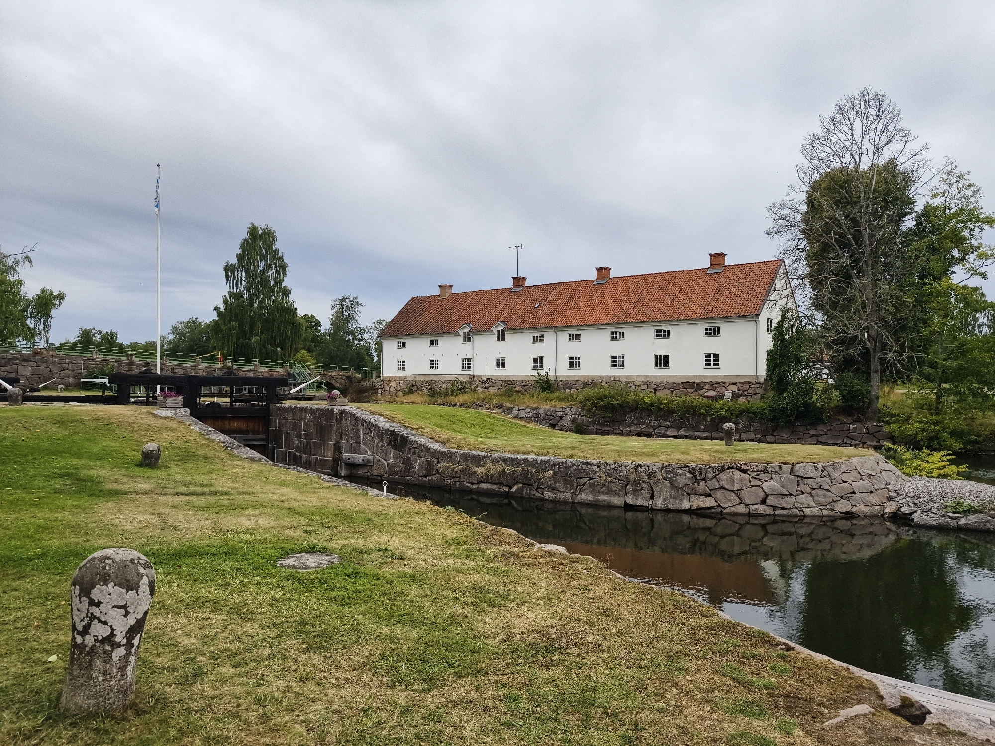 The image shows a canal lock and a white, two-story building with a red tile roof in Brokind, Sweden. A white flagpole with a blue and white flag is visible on the left side of the frame, near a stone bridge crossing the canal. The building, which appears to be an old manor or administrative building, has multiple windows with white frames and red shutters. The canal walls are constructed of rough-hewn stones. Grassy areas surround the canal and the building. Trees, some with full green foliage and one bare, are scattered throughout the scene. The sky is overcast with grey clouds. Two weathered stone posts are present in the grassy area in the foreground. The water in the canal is dark and reflects the overcast sky.