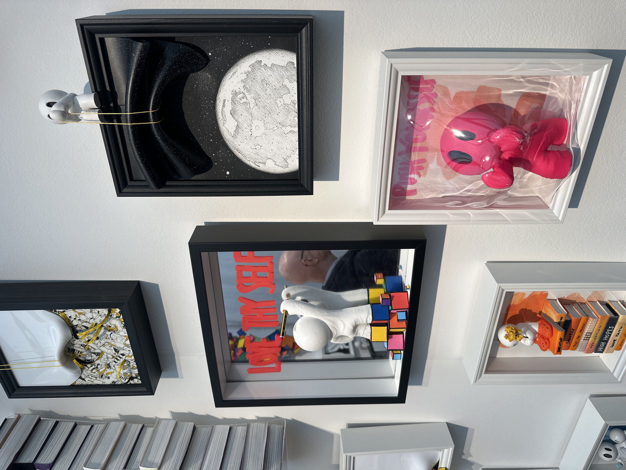 The image shows a collection of framed artworks displayed on a white wall, likely inside an art gallery.
The top-left artwork features a black void and a drawn moon, seemingly connected to a white figure by a yellow cord. The top-right artwork contains a pink, toy-like figure encased in plastic wrap.
The central artwork presents a mirrored image, reflecting a person holding a pencil to a white ball, positioned amidst colorful blocks and the words "LOVE THY SELF".
The bottom-left artwork shows a white figure and a background of drawings connected by a yellow cord. The bottom-right artwork displays miniature books with titles like "BREATHE", "SUCCESS", "HIGH HOPES", and "HAVE FAITH", along with a small figure covered in yellow petals.
Based on the style of art and display, the location could be within an art gallery in London, United Kingdom.
