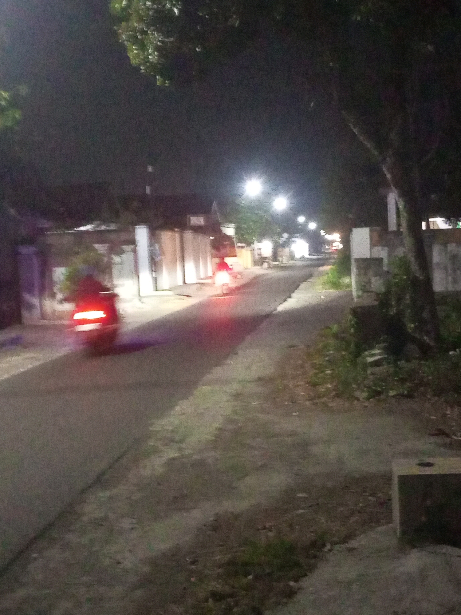 This image depicts a street scene at night in Ngasem, Indonesia. A person is riding a motorcycle on a paved road, with the motorcycle's taillight illuminated. The road is flanked by a sidewalk with some vegetation and a tree. In the background, there are buildings with lights on, suggesting a residential or commercial area. Streetlights are also visible, illuminating parts of the street. The overall impression is that of a quiet, dimly lit street in a town or village.