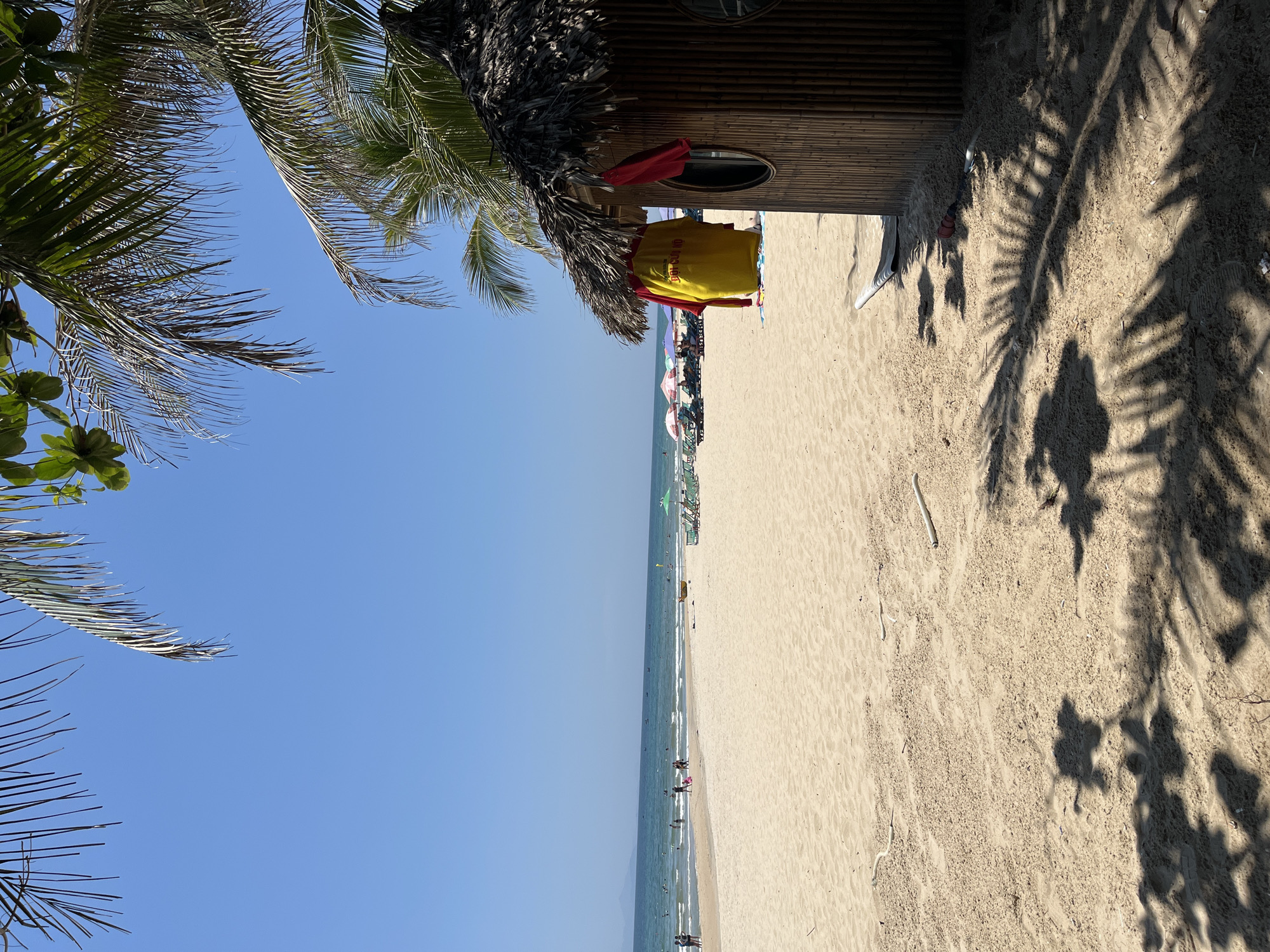 The image depicts a beach scene in Đà Nẵng, Vietnam, taken from an elevated position. A lifeguard station, constructed from bamboo and topped with a thatched roof, stands to the right, with a red and yellow lifeguard uniform hanging on the exterior. A sandy beach stretches out, populated with beachgoers, umbrellas, and chairs in the distance. The ocean meets the clear, blue sky at the horizon. Palm trees frame the left side of the image, and shadows of palm leaves are cast across the sand.