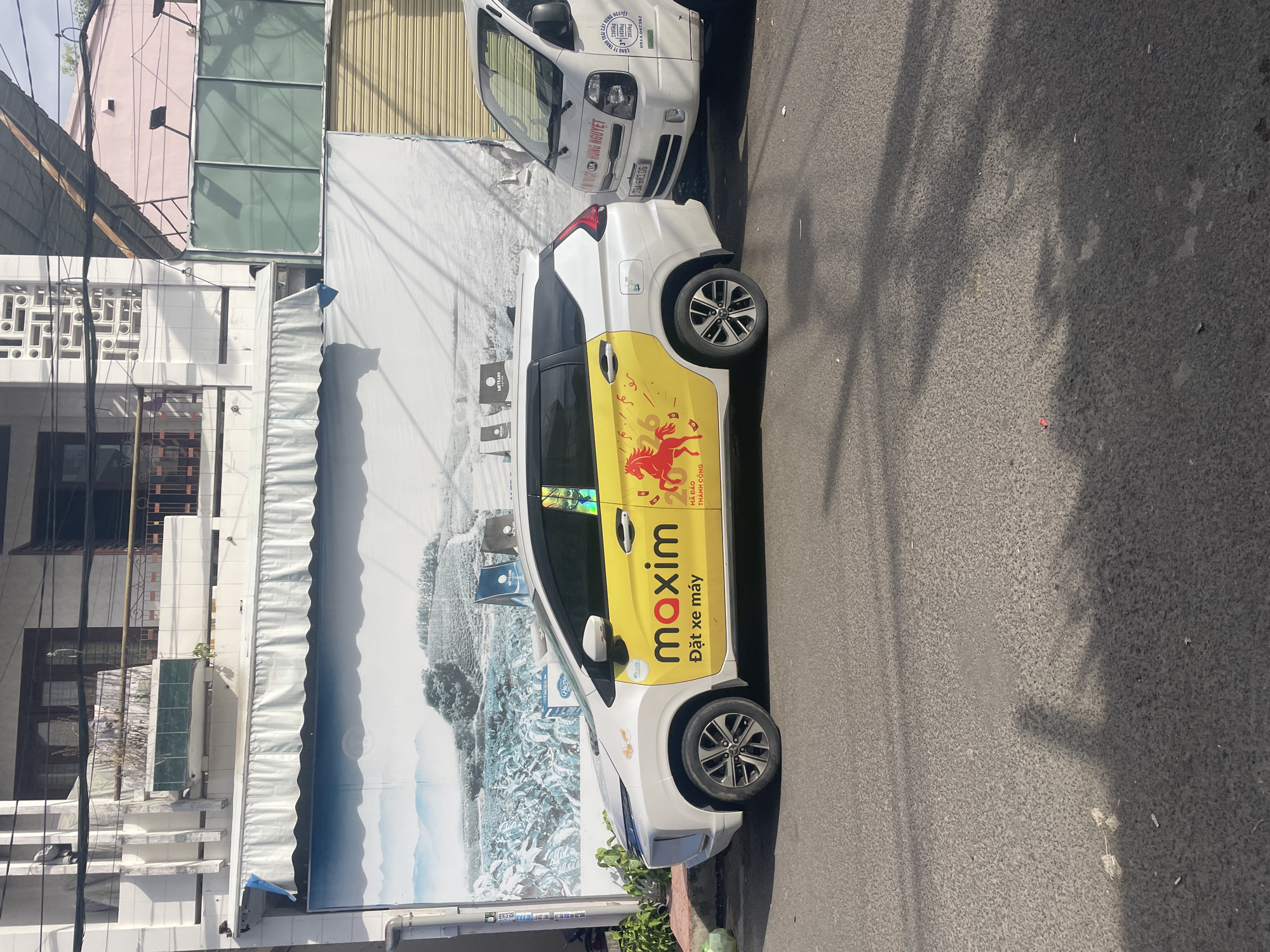 The image depicts a street scene in Nha Trang, Vietnam. In the foreground, a white car with a yellow stripe and the "maxim" logo is parked on the asphalt road. The yellow stripe features a red horse graphic and Vietnamese text. To the right of the car, a white truck is parked. In the background, buildings with awnings and a large mural are visible. Overhead power lines crisscross the scene. The lighting suggests it is daytime, with shadows cast on the road.