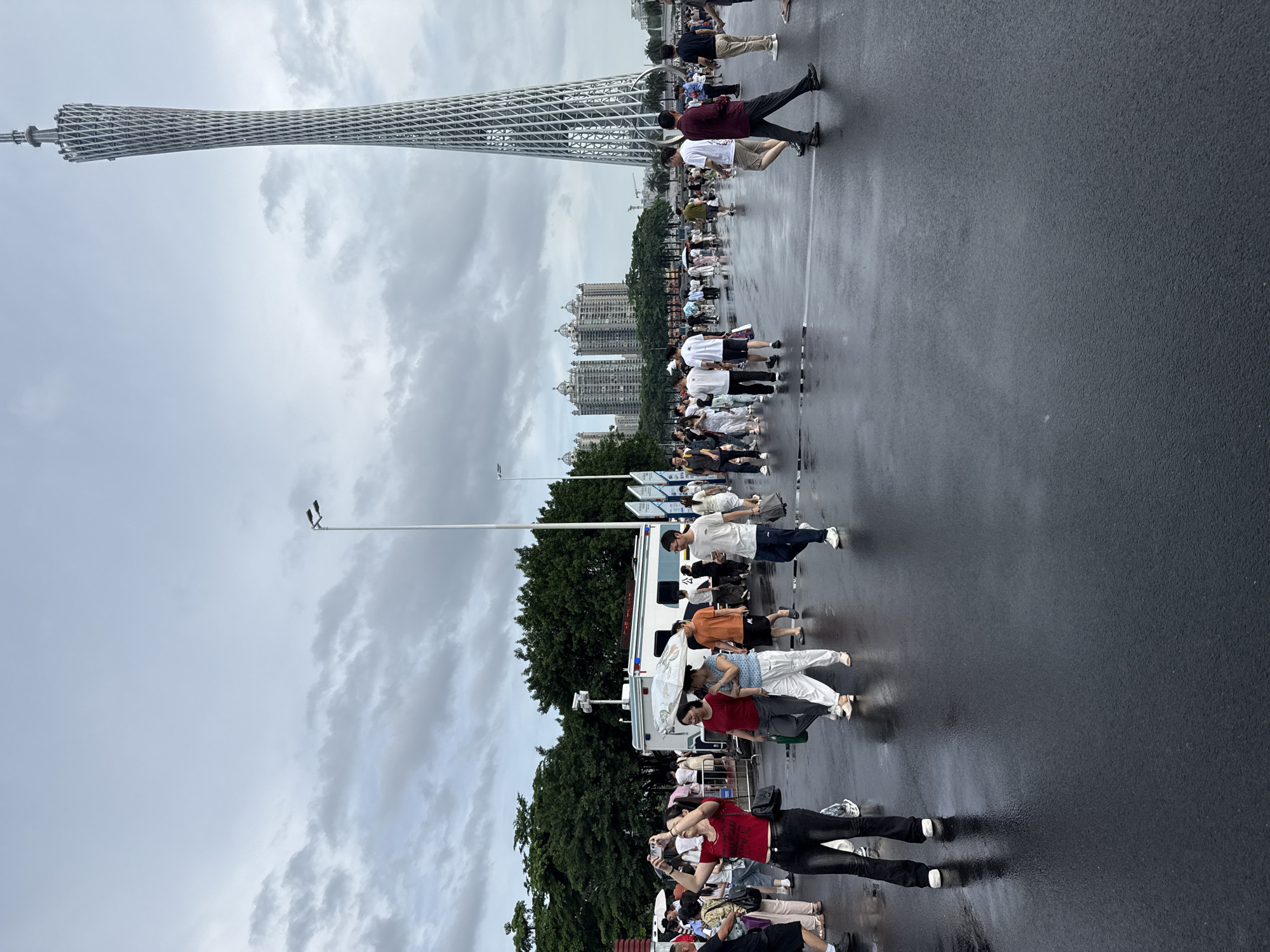 The image depicts a street scene in Tianhe District, China, with the Canton Tower prominently featured in the background. A crowd of people are walking on a wet, paved surface, indicating it may have recently rained. Several individuals are holding umbrellas, reinforcing this observation. In the foreground, a woman in a red top and black pants is taking a photo with her phone. To her left, another woman in a white top and patterned skirt is also engaged with her phone. Further back, a white van with a security camera mounted on top is parked. The sky is overcast with heavy clouds. Buildings and trees are visible behind the crowd.