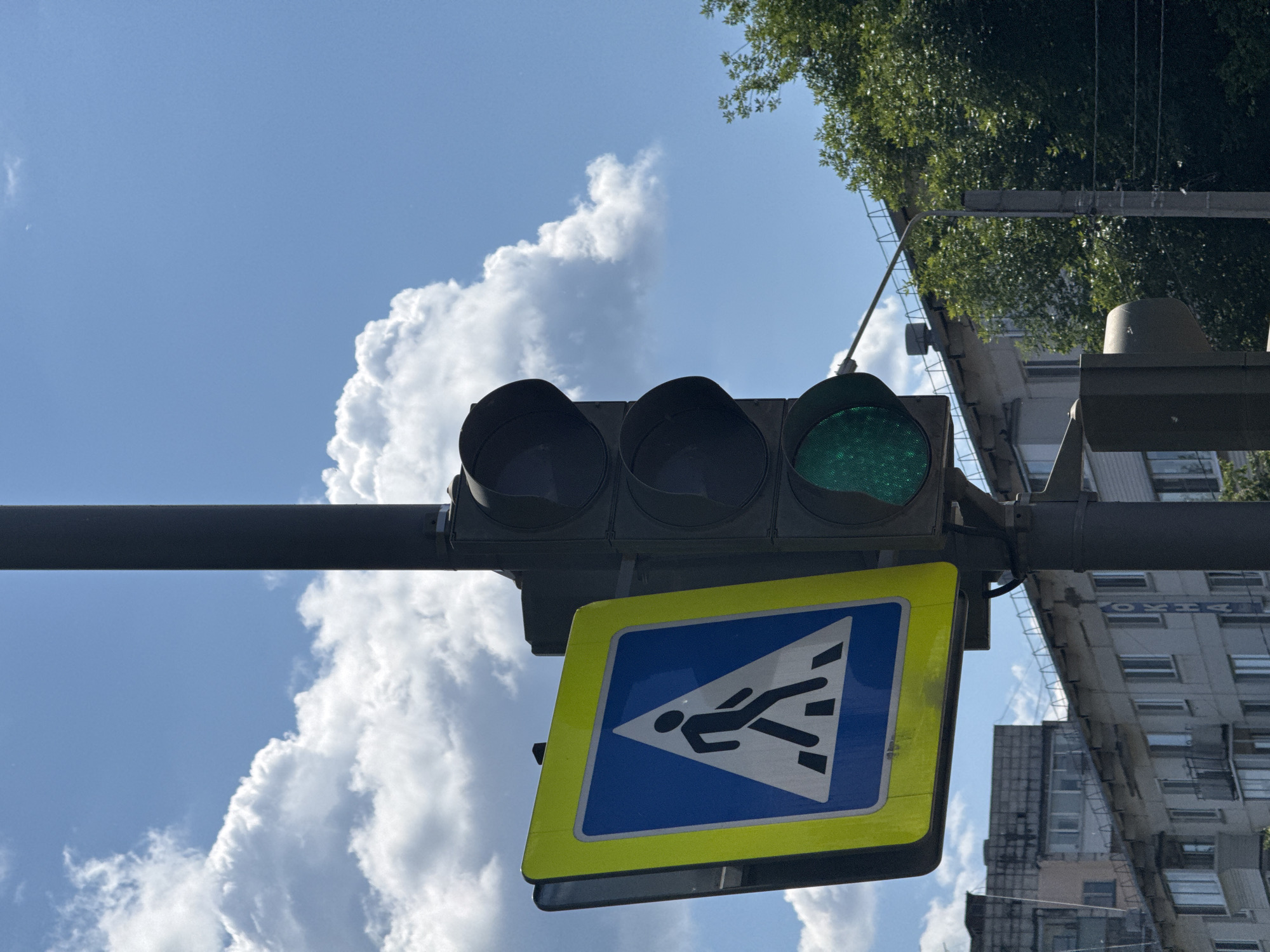An outdoor, eye-level, medium shot captures a traffic light pole with a pedestrian crossing sign on a bright, cloudy day. The pedestrian crossing sign, a blue square with a white triangle containing a black silhouette of a person walking across stripes, is mounted to the left of the traffic light. The traffic light itself is dark, with three large, circular lenses stacked vertically. The bottom lens is illuminated green, indicating that pedestrians may cross. Behind the traffic light pole and sign, a line of apartment buildings stretches across the background. Lush green trees can be seen to the right of the frame. The sky is a vibrant blue, dotted with fluffy white clouds. The scene appears to be a typical urban streetscape in a temperate climate.