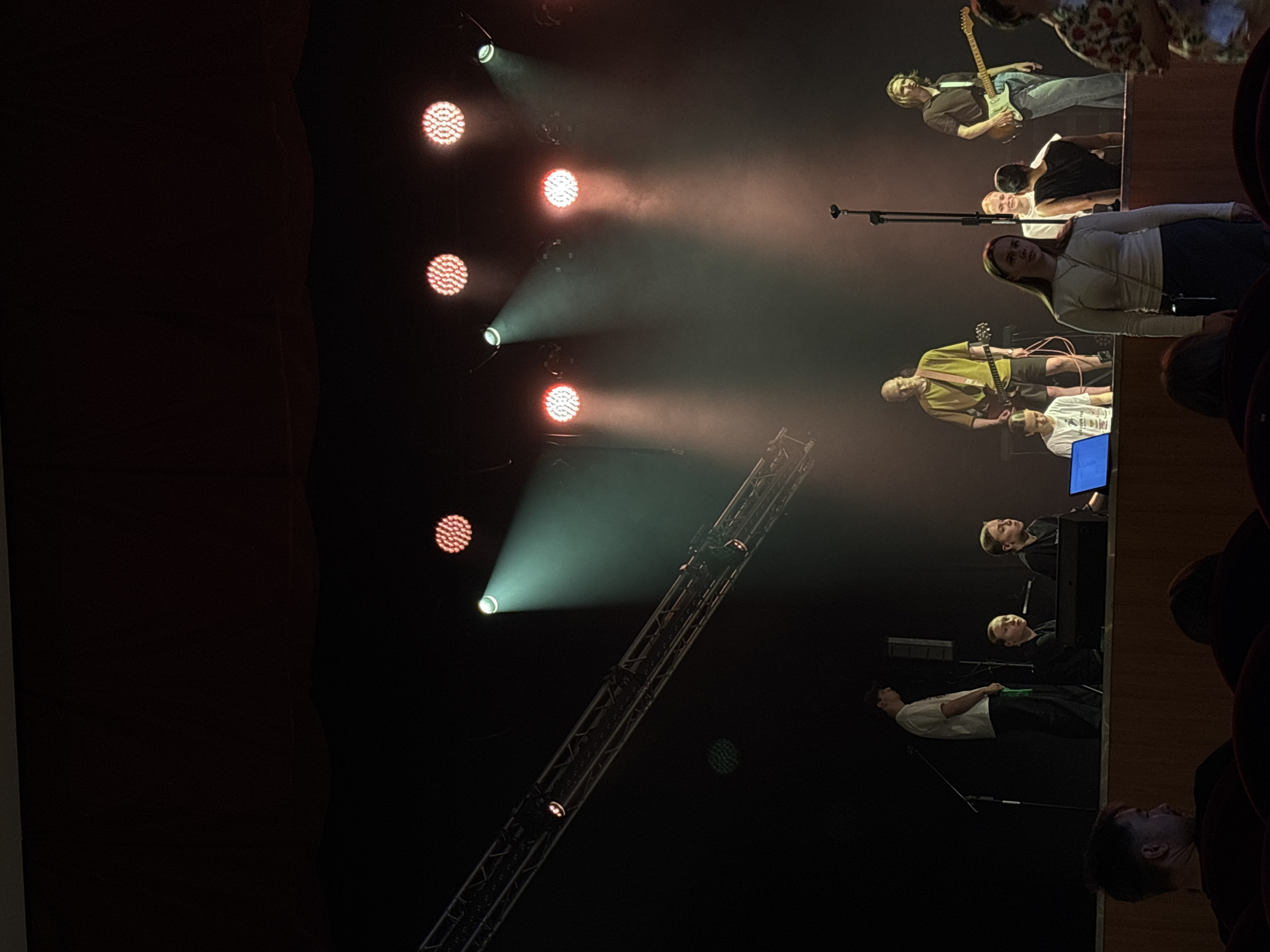 A group of young people are on a stage, performing. Some are playing instruments, like guitars, and others appear to be singing or listening. Stage lights illuminate the performers and create beams of light through the smoky air. The scene suggests a concert or live performance. The individuals are casually dressed, with one person wearing a yellow shirt and another a black long-sleeved shirt. The backdrop is dark, with some stage equipment visible, including a lighting rig. The overall mood is energetic and focused on the performance. The audience is not visible, but the seating in the foreground suggests a theater or concert hall. The time of day is likely evening or night, given the artificial lighting and concert setting.