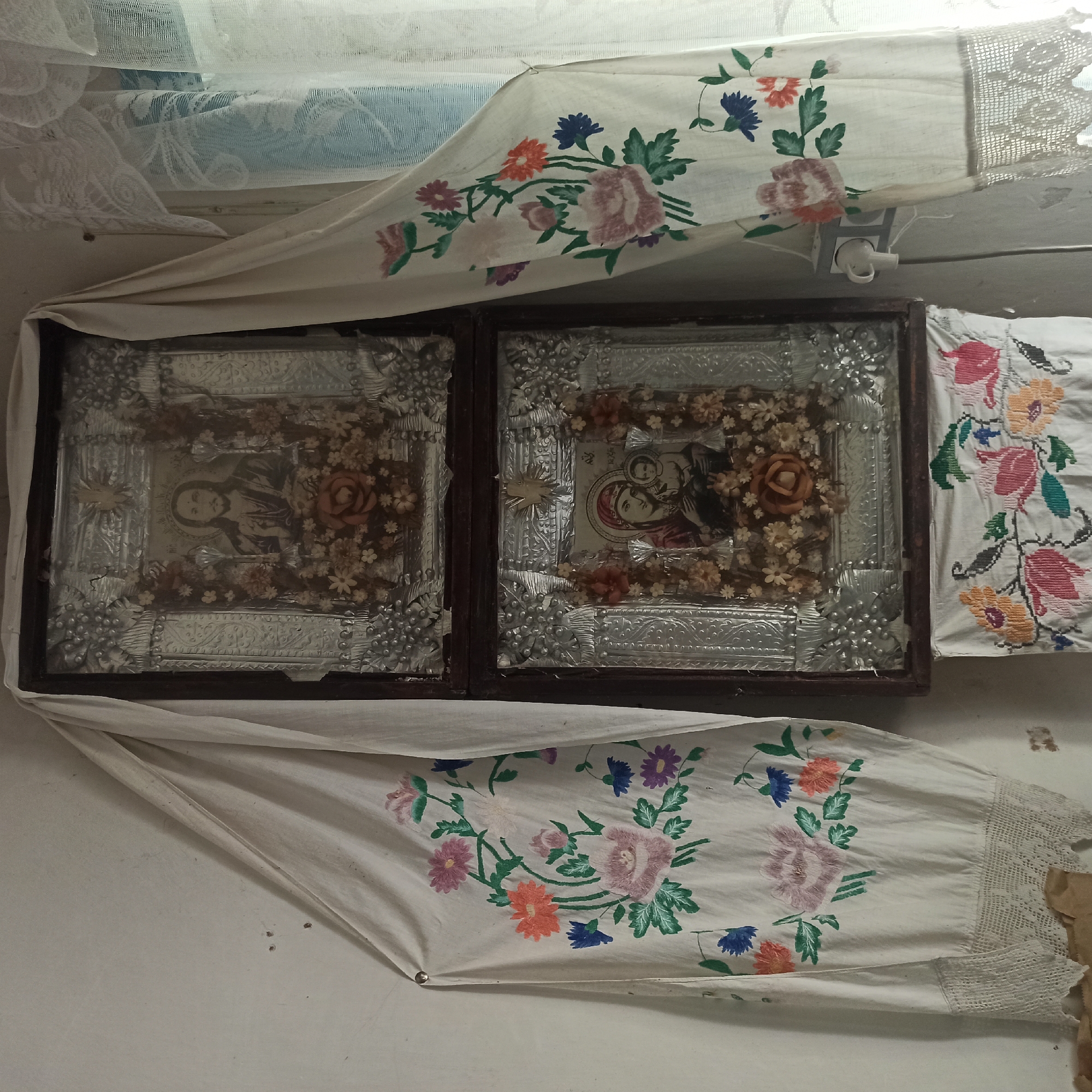 The image shows two icons hanging on a white wall. The top icon depicts Jesus Christ, and the bottom icon shows the Virgin Mary holding the infant Jesus. Both icons are framed and covered with decorative metalwork and dried flowers. White embroidered cloths with floral patterns hang on either side of the icons, framing them. A piece of embroidered fabric with a floral pattern is also visible hanging below the bottom icon. A white curtain with lace trim is visible to the right of the icons. The wall appears to be plaster and shows some signs of wear. There is a light switch and an electrical outlet on the wall. The lighting in the scene is soft, suggesting indoor lighting or diffuse natural light coming from the window. The overall impression is of a domestic or religious space.