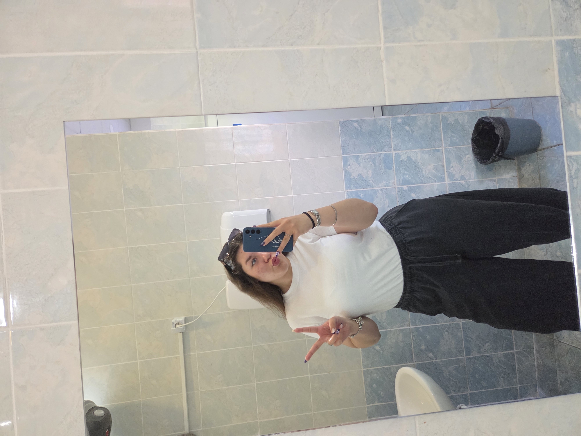 The image shows a young woman taking a selfie in a bathroom. She is standing in front of a tiled mirror, wearing a white t-shirt and dark pants. She is holding a blue smartphone and making a peace sign with her hand. Sunglasses are perched on her head. In the reflection, the bathroom features light blue tiled walls and floor. A waste bin, a sink, and a toilet paper dispenser are visible. The specific location in Stara Zagora, Bulgaria, cannot be determined from the image alone.