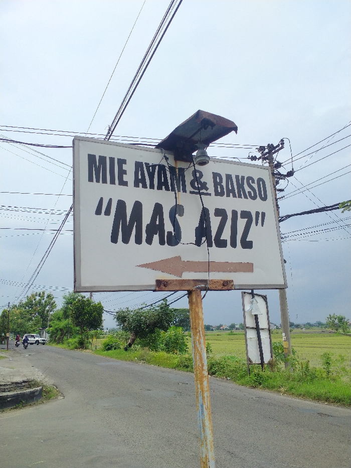 This media file depicts a rural or semi-rural roadside scene in Kecamatan Kebakkramat, Indonesia, under an overcast sky. In the foreground, a prominent white rectangular sign, supported by a rusty metal pole, directs travelers. The sign displays black text reading "MIE AYAM & BAKSO" (Indonesian for Chicken Noodles & Meatballs) and ""MAS AZIZ"", along with a brown arrow pointing to the left. A small solar panel and a light fixture are mounted above the sign, likely for nighttime illumination.
The setting includes a paved road with green trees and bushes lining its side. Beyond the vegetation, extensive green rice fields (paddy fields) are visible, extending into the distance where some scattered buildings can be discerned. Numerous electrical wires crisscross the sky, connected to utility poles, one of which stands on the right side of the road.
On the road, a white car and a scooter carrying at least one person are seen, indicating general traffic activity. Another individual is visible near the car. A second, partially visible billboard or sign structure stands further down the road on the right. The scene conveys a sense of local life, with the sign advertising a modest food establishment in an agricultural landscape. The weather is cloudy, providing soft, diffused light during the daytime.