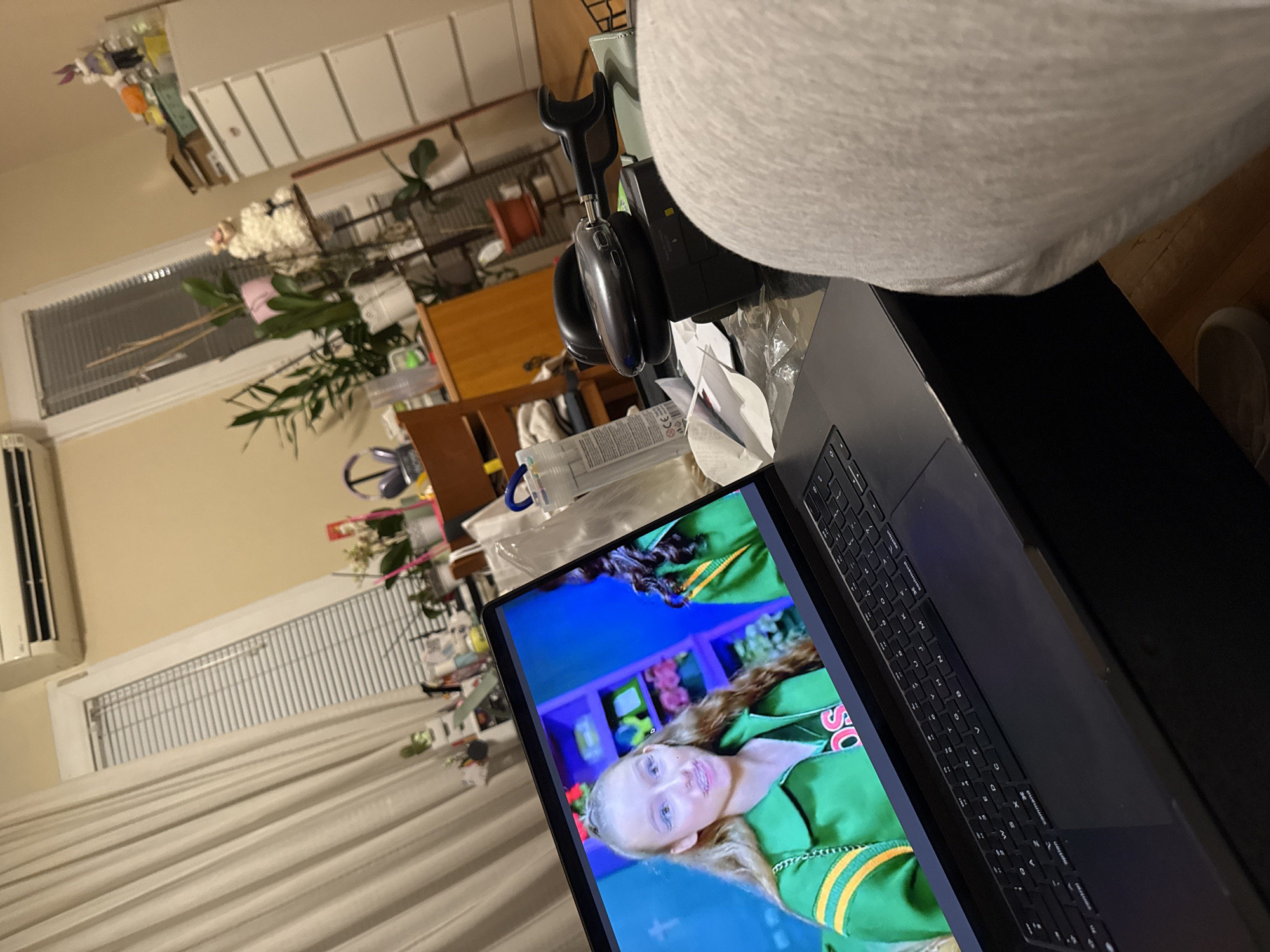 The image shows a room with various objects. A laptop is in the foreground, displaying a person in a green outfit on the screen. To the left of the laptop are headphones and various containers on a wooden table. In the background are windows with blinds, plants, and storage units. An air conditioning unit is visible on the wall. The location is indiscernible from the provided image.