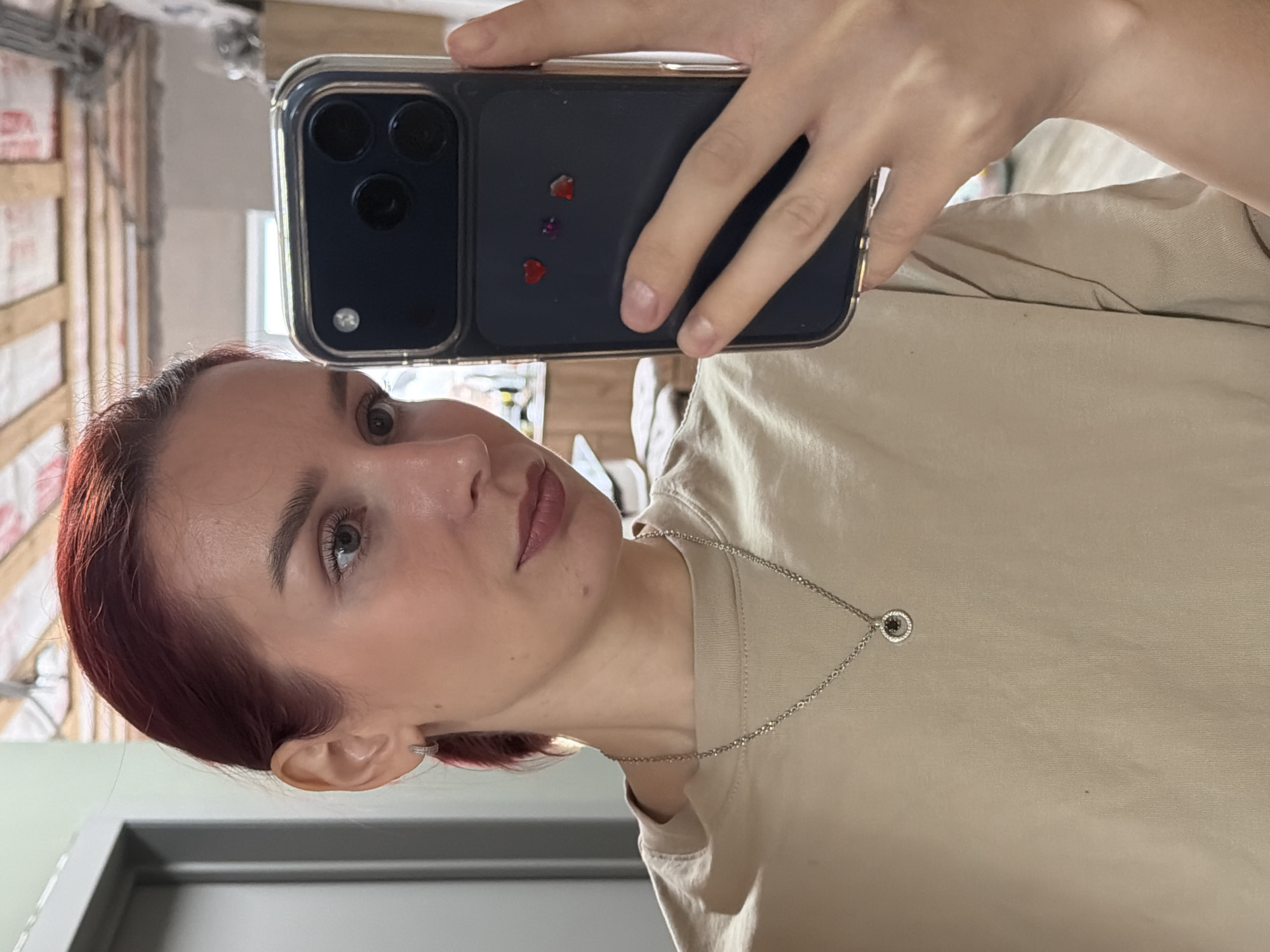 A woman is taking a selfie with a smartphone. She has red hair and is wearing a beige t-shirt and a silver necklace. The smartphone has a dark blue case with heart-shaped decorations. The background is out of focus, but it appears to be a room with light green and grey walls. The location is likely Chaikovsky, Russia, although this is not explicitly clear from the image.