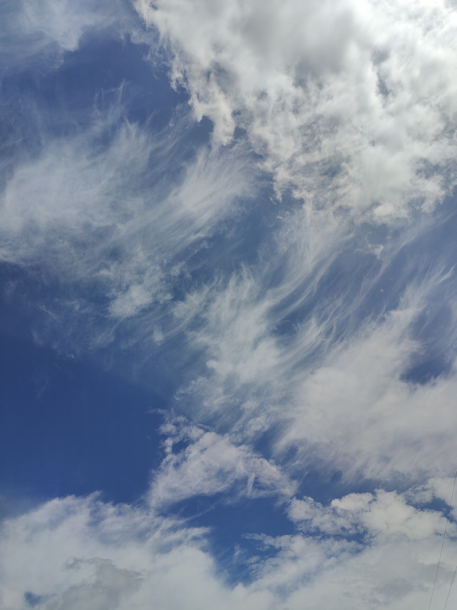 The image shows a wide-angle view of a sky with scattered clouds. The sky is a deep blue, with wispy white clouds that appear to be cirrus clouds, streaking across the frame. There are also larger, puffier clouds in the upper right portion of the image. The lighting suggests it is daytime, possibly late morning or early afternoon, with the sun illuminating the clouds from the top. The scene is natural, with no man-made structures or people visible. The image is taken from a low angle, looking upwards. The location is identified as Kryvyi Rih, Ukraine.