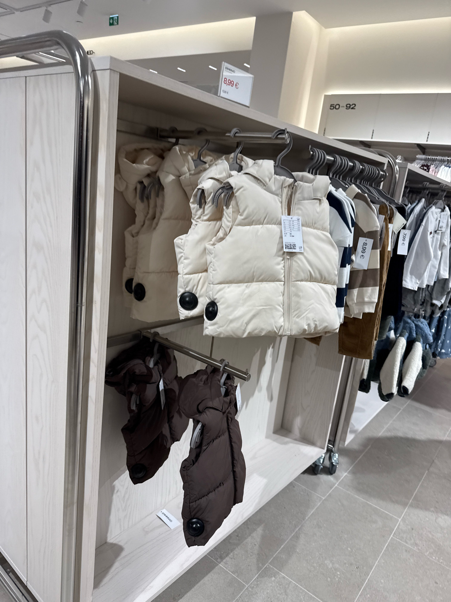 The image shows a retail store interior in Tallinn, Estonia, specifically within a clothing store. The focus is on a display of children's outerwear, featuring several cream-colored puffer vests and some brown puffer jackets. The garments are neatly hung on hangers on a metal rack system. Some of the vests have price tags with the cost indicated as 8.99 €. To the right of the vests, other children's clothing items like striped long-sleeved shirts and white long-sleeved tops are visible, also on hangers. A sign overhead reads "50-92", indicating the size range of the clothing. The store appears modern and well-lit, with light-colored wooden shelving units and a grey tiled floor. There are no people visible in the frame. The overall impression is of a clean and organized retail environment.