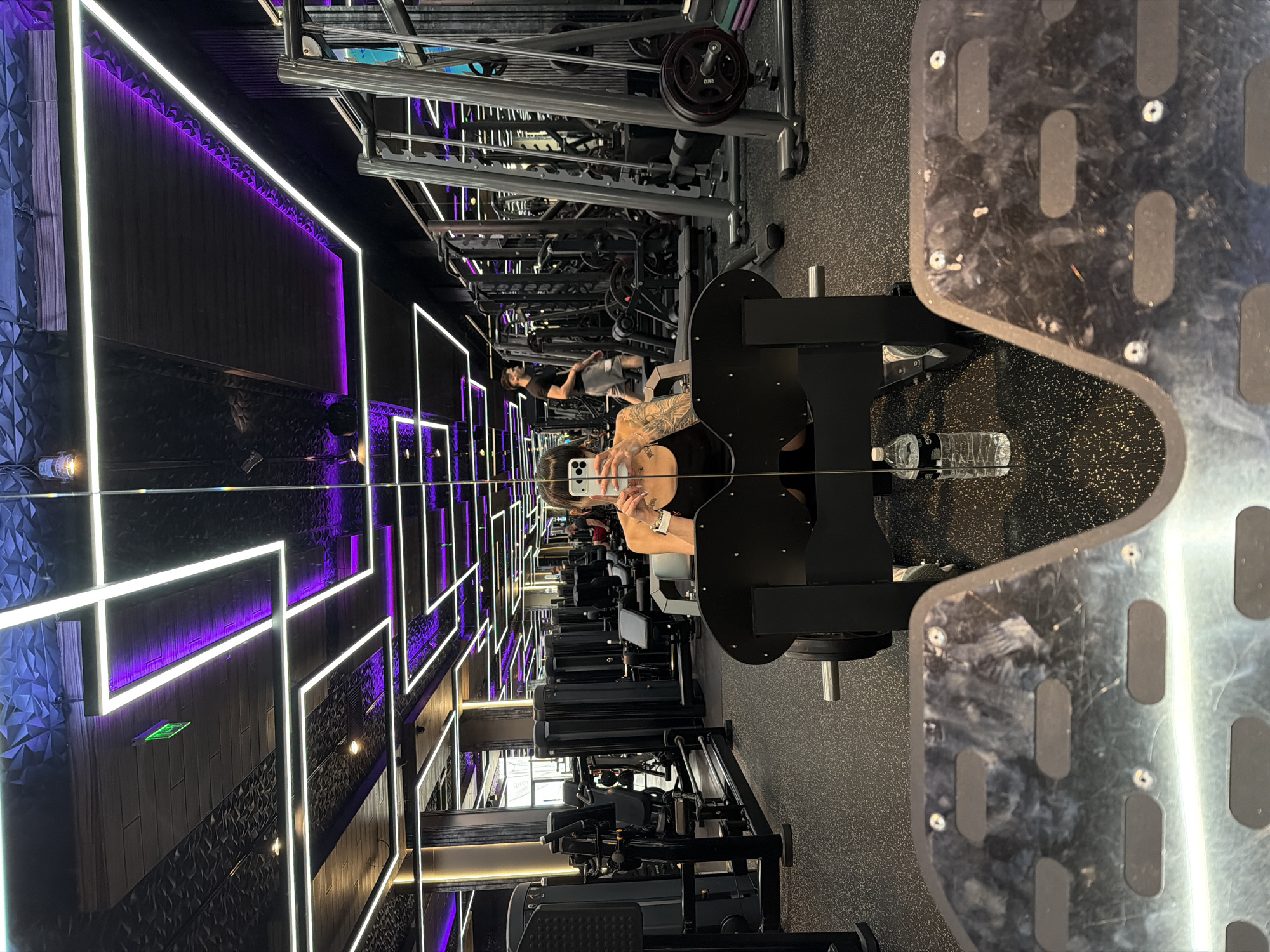 The image shows a woman taking a selfie in a modern, well-lit gym. The gym features black equipment with silver accents and a black, textured ceiling with integrated purple and white LED lighting. The floor is black rubber with speckled grey particles. In the background, several rows of gym machines and other individuals can be seen. The woman is wearing a black tank top and appears to have tattoos on her arm. A water bottle is placed on the floor next to the gym equipment she is using. There is no information in the image to indicate the location is in Plovdiv, Bulgaria.