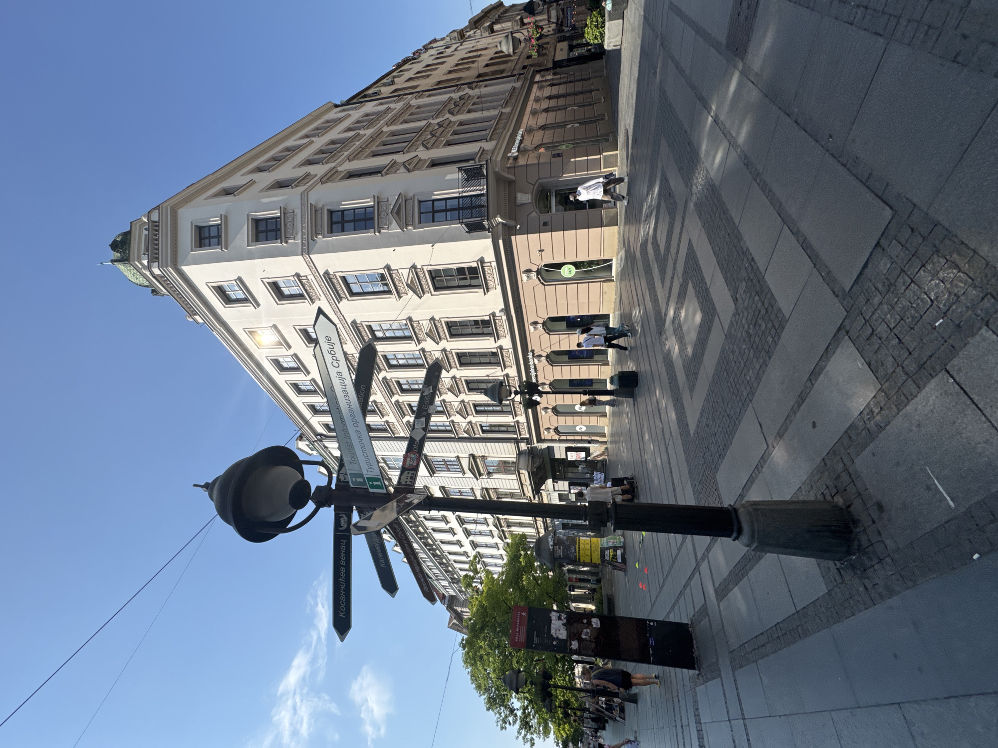 The image shows a street scene in Belgrade, Serbia. In the foreground, a lamppost with multiple directional signs stands prominently. The signs point to various locations, including "Kosancicev venac," "Kalemegdan," and "Turisticka organizacija Srbije" (Tourist Organization of Serbia). The street is paved with large stone tiles and smaller cobblestones, forming a geometric pattern.
Several people are walking on the pedestrianized street. On the left, a woman in a dark top and shorts walks away from the camera, while another person walks towards the camera. Further down the street, more individuals are visible, some dressed in casual attire.
To the right of the lamppost, a large, multi-story building with a cream-colored facade and green domed roof dominates the background. The building features numerous windows and ornate architectural details. Shops are visible at street level, with signs such as "NLB Komercijalna banka" and "24h."
On the left side of the street, a row of trees provides shade, and a black, vertical information board is placed near the lamppost. The sky is clear and blue, indicating a sunny day. The overall scene depicts a busy urban environment in a European city.