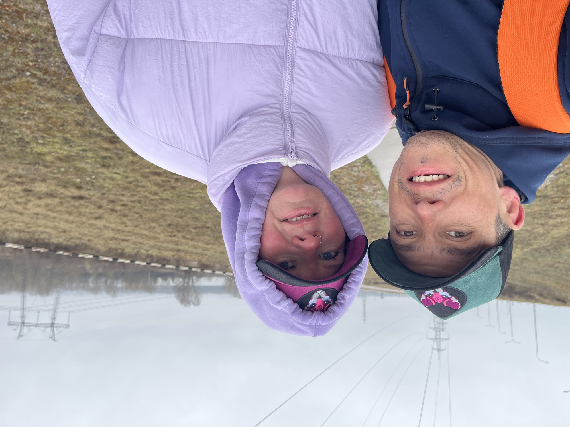 This close-up selfie-style image features two individuals, a woman and a man, smiling brightly at the camera in an outdoor setting. The image is presented rotated 90 degrees counter-clockwise.
The woman, positioned on the left, wears a light lavender padded jacket with the hood up, covering her hair. She also has a dark pink and black baseball cap underneath her hood, featuring a graphic of a creature, possibly a pig, with prominent ears and snout. She has a small septum piercing and a wide smile.
The man, on the right, wears a navy blue jacket with a contrasting orange strap or panel visible on his right shoulder. He has a dark green and black baseball cap, also featuring a graphic similar in style to the woman's cap, which appears to be a creature. He displays a broad smile, revealing his teeth, and has short stubble.
The background, largely out of focus, shows a somewhat desolate landscape. On the left (which would be the bottom when correctly oriented), there is brownish-yellow ground with sparse vegetation, suggesting either late autumn or early spring. A faint barrier or guardrail runs horizontally across the middle distance. The sky is overcast and light gray, indicating a cloudy day. In the distance, several tall power line pylons and power lines are visible, extending across the scene, particularly prominent on the right side (top when correctly oriented). Bare trees or bushes can also be seen faintly on the horizon.
The overall mood conveyed by the subjects' smiles is cheerful and friendly. The scene suggests a location in a rural or semi-rural area, possibly alongside a road or path, fitting the provided context of Chaikovsky, Russia. No visible text is present in the image.