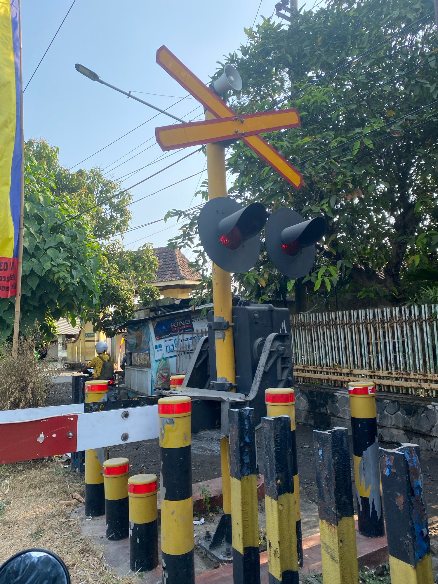 This is a full shot of a railroad crossing in Rembang, Indonesia, on a sunny day. A yellow pole with a red and yellow crossbar dominates the center of the frame. Two red lights are visible on the crossbar, indicating the crossing is active. In the background, a small stand with a banner is visible. A person on a motorcycle wears a yellow helmet and vest. A tall fence made of white poles stretches across the background. The ground is covered in dry grass and dirt. The image captures a typical scene at an Indonesian railroad crossing, likely in the city of Rembang.