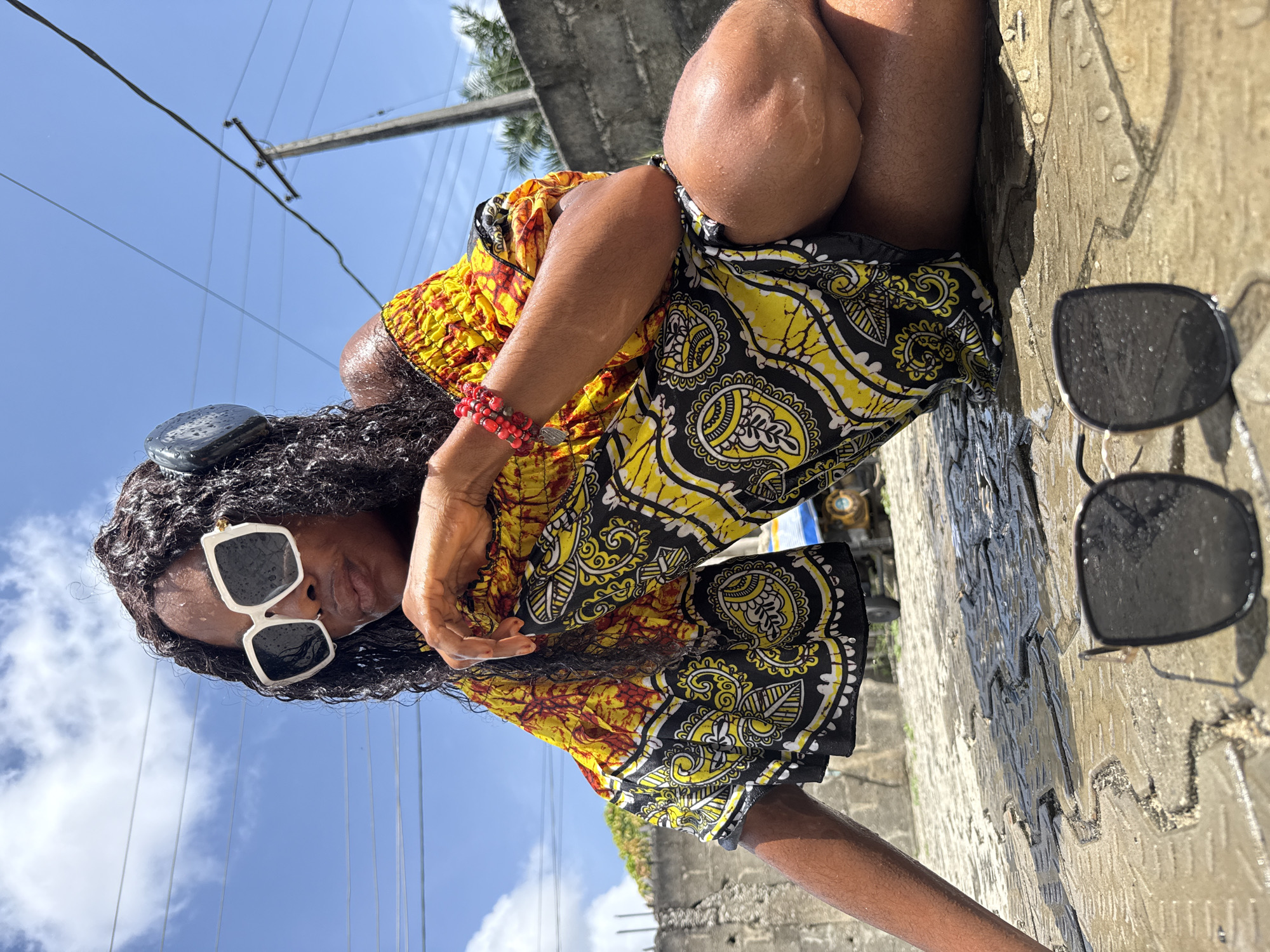 A photograph taken in Ado, Nigeria, shows a Black woman in a patterned yellow and black dress seated outdoors. She is wearing white sunglasses and a red beaded bracelet. Her hair is long and dark. The woman's skin appears wet. Next to her on the ground, two pairs of sunglasses are visible. The background includes a concrete wall, a blue sky with clouds, and utility poles with wires. The ground is wet and textured.