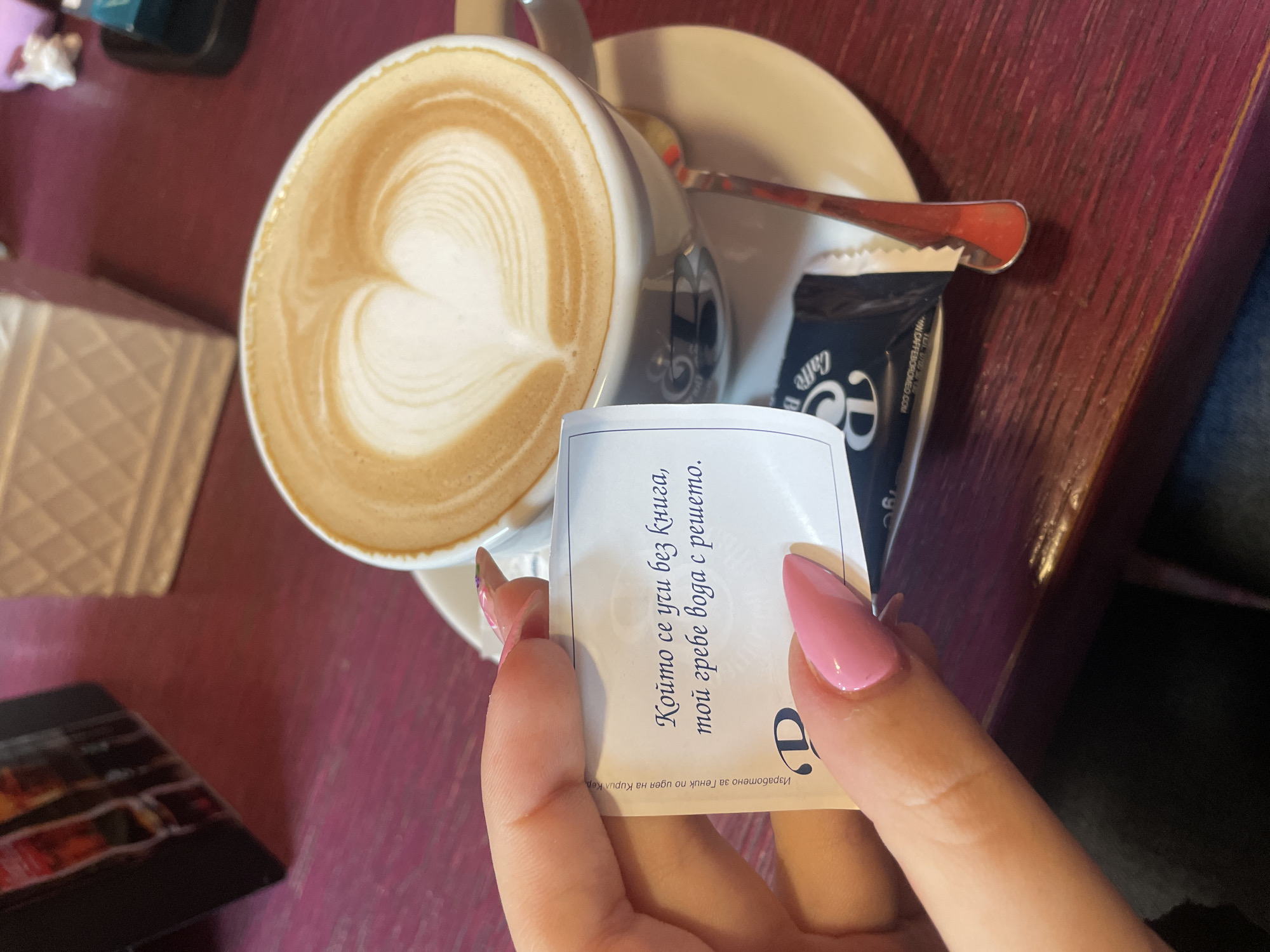 The image shows a cup of coffee with latte art, a sugar packet, and a hand holding a small paper slip with Bulgarian text. The coffee is in a white cup and saucer, with a spoon resting on the saucer. The sugar packet is black and white with the word "Caffe" on it. The paper slip has Bulgarian text and a "D" logo. The hand has long, pink nails. The background includes a red wooden table. The text on the paper slip suggests the image was taken in Bulgaria. The sugar packet shows "CAFFEBOROMEO.COM" which leads to a search result for Cafe Borromeo in Varna, Bulgaria.