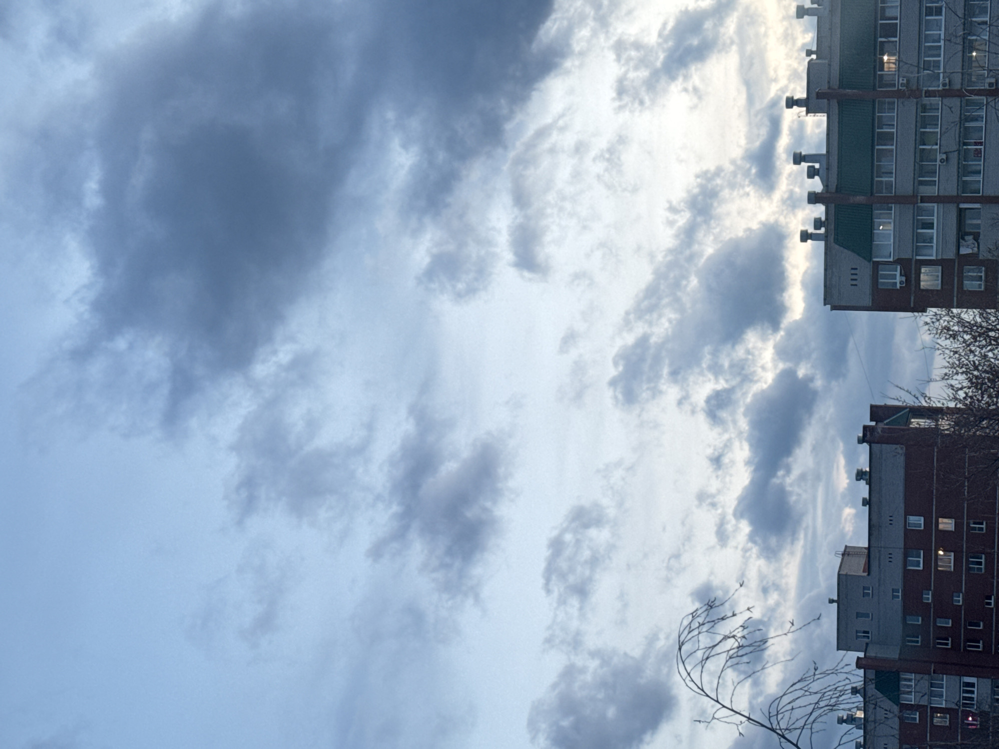 The image, presented rotated counter-clockwise, captures an urban sky with multi-story residential buildings and bare tree branches. The sky is predominantly overcast, displaying a mix of dark grey clouds and brighter, lighter blue patches where sunlight filters through.
Two distinct apartment blocks are visible on what would be the right side of a correctly oriented image. The upper building features a grey and white facade with sections of green roofing and several cylindrical structures, possibly vents or chimneys, protruding from its roofline. Numerous windows are visible, some reflecting the sky, others showing interior darkness, and at least one with an interior light on. The lower building, partially constructed with red brick and partly with a lighter grey material, also has many windows, a few of which display illuminated interiors.
In the foreground, particularly in the lower right, are the delicate, leafless branches of a tree, suggesting a season like late autumn, winter, or early spring. The overall lighting indicates daytime, despite the cloudy conditions and some illuminated windows. No people or specific activities are depicted. The architectural style of the buildings is consistent with residential areas found in Russia. No visible text is present in the image. The scene conveys a quiet, typical urban setting under a cloudy sky.
