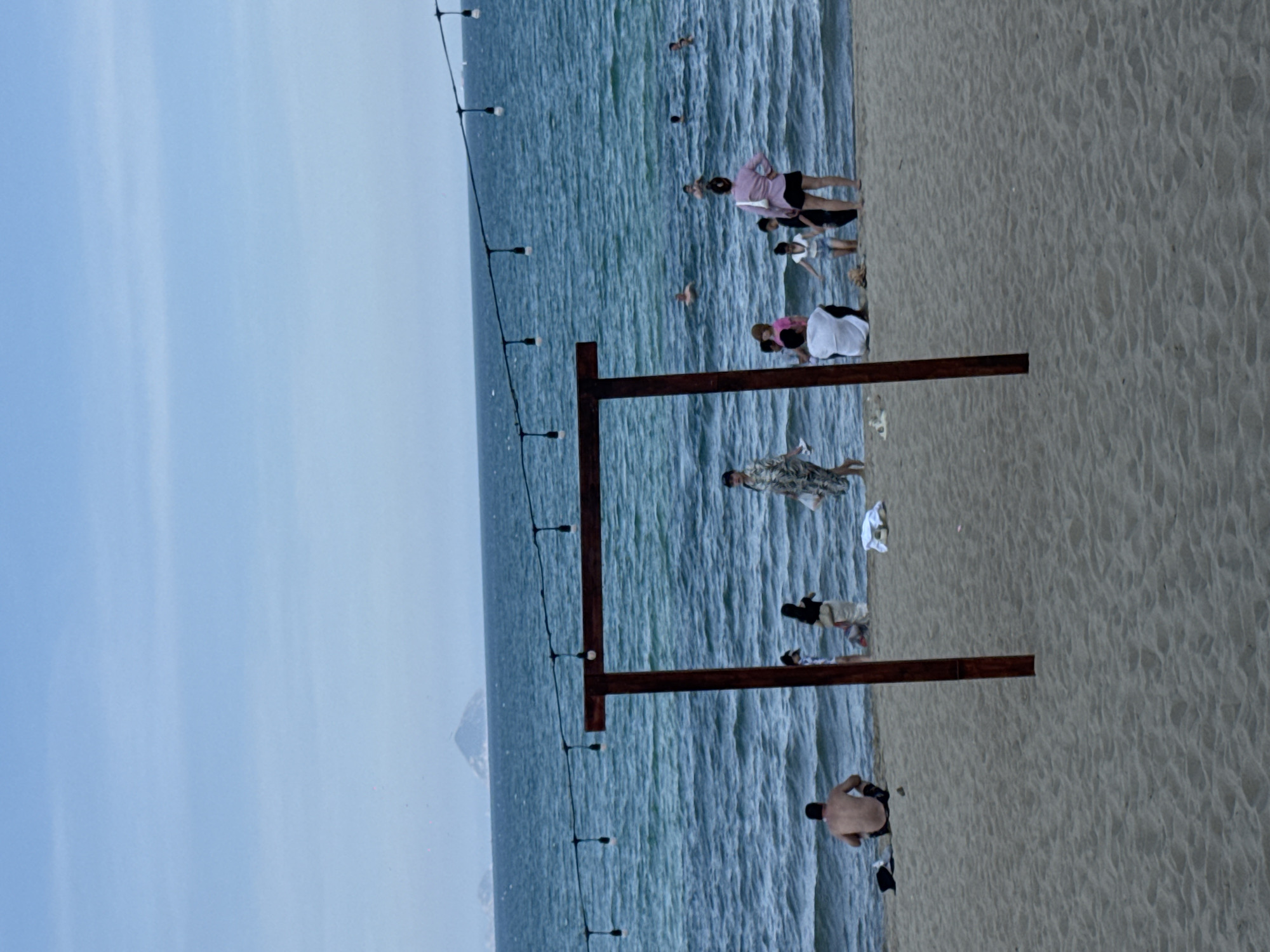 The image depicts a beach scene in Nha Trang Ward, Vietnam, under a cloudy sky. In the foreground, a wooden archway stands on the sand, with a string of lights overhead. Several people are visible on the beach and in the water. On the left, a shirtless man sits on the sand facing the ocean. Further back, a group of individuals, including a woman in a floral dress and others in casual beach attire, are gathered near the water's edge. Some people are wading or swimming in the ocean. The sea is a vibrant blue-green, with moderate waves. In the distance, a hazy island or landmass is visible on the horizon. The beach itself is sandy, with some scattered debris and towels.
