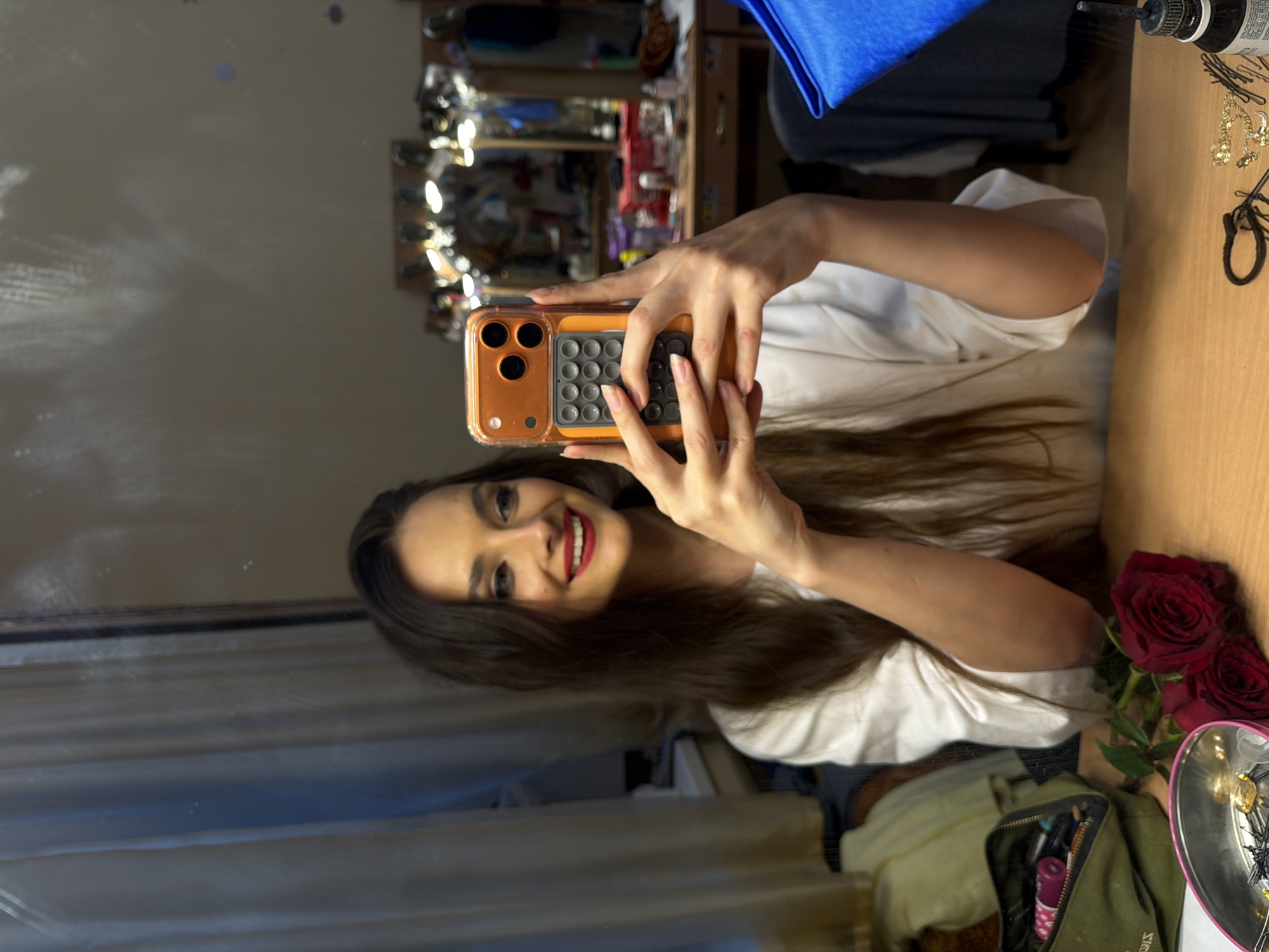 In an indoor scene, a young woman with long, dark hair and bright red lipstick is taking a selfie in a mirror. She is wearing a white t-shirt and holding an orange-cased smartphone in her hands. The background shows a vanity area with lights and mirrors, suggesting a dressing room or backstage area. On the table in front of her, there are two red roses, a green makeup bag, and various hair accessories. The lighting is warm, with reflections visible in the mirror.