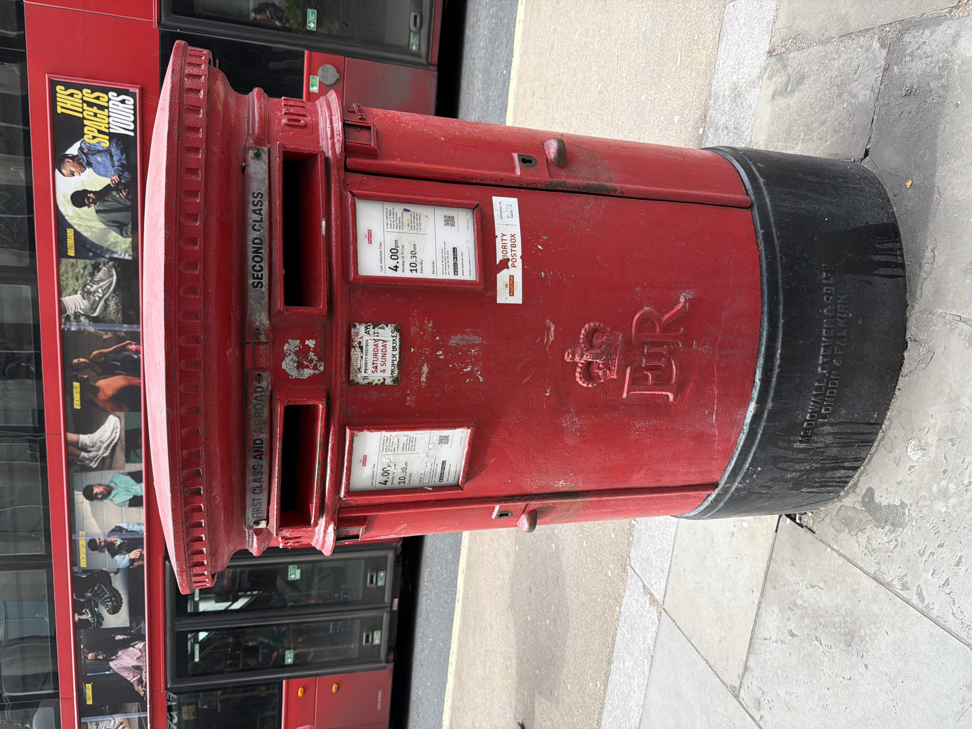 The image shows a red cylindrical Royal Mail postbox on a paved sidewalk in London, UK. The postbox features separate slots for "First Class and Abroad" and "Second Class" mail. It also displays a collection schedule indicating a last collection time of 4:00 pm on weekdays and 10:30 am on Saturdays. The postbox is marked with the Royal Cypher "ER" beneath a crown. "MEDOWALL STEVENAGOITU & FALKIAN" is imprinted on the lower part of the postbox.
A red double-decker bus, marked with an advertisement, is parked beside the postbox. The bus advertisement is a visual campaign showing people's faces and bodies wearing clothes and shoes.