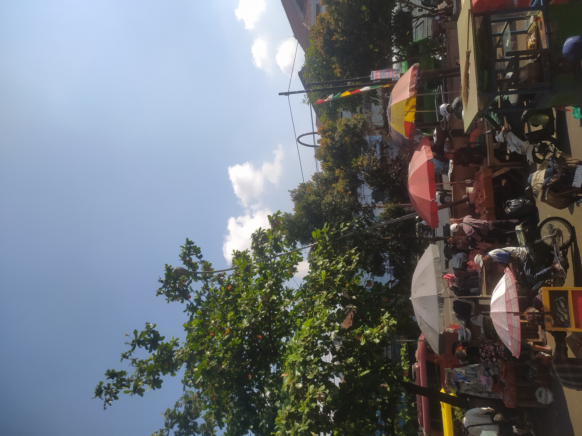The image depicts an outdoor market scene under a clear blue sky with scattered clouds. A large tree with green leaves and some reddish-orange fruit is prominent in the foreground on the left, casting shade. Several market stalls are visible, covered by colorful umbrellas, including red, white, and yellow ones. People are browsing and interacting at the stalls, which appear to be selling various goods, possibly including food and textiles.
In the background, there are trees, buildings with red tile roofs, and a traffic light. A few vehicles, including a red bus or truck and motorcycles, are present, suggesting a public road or thoroughfare adjacent to the market. A flagpole with a tricolor flag (red, white, and green) is also visible. One person is riding a motorcycle, and another is standing near a stall with a broom on display. The overall impression is a lively daytime market atmosphere. Based on the presence of Indonesian flags and the general appearance of the market and surrounding architecture, this scene is likely located in Kupang, Indonesia.