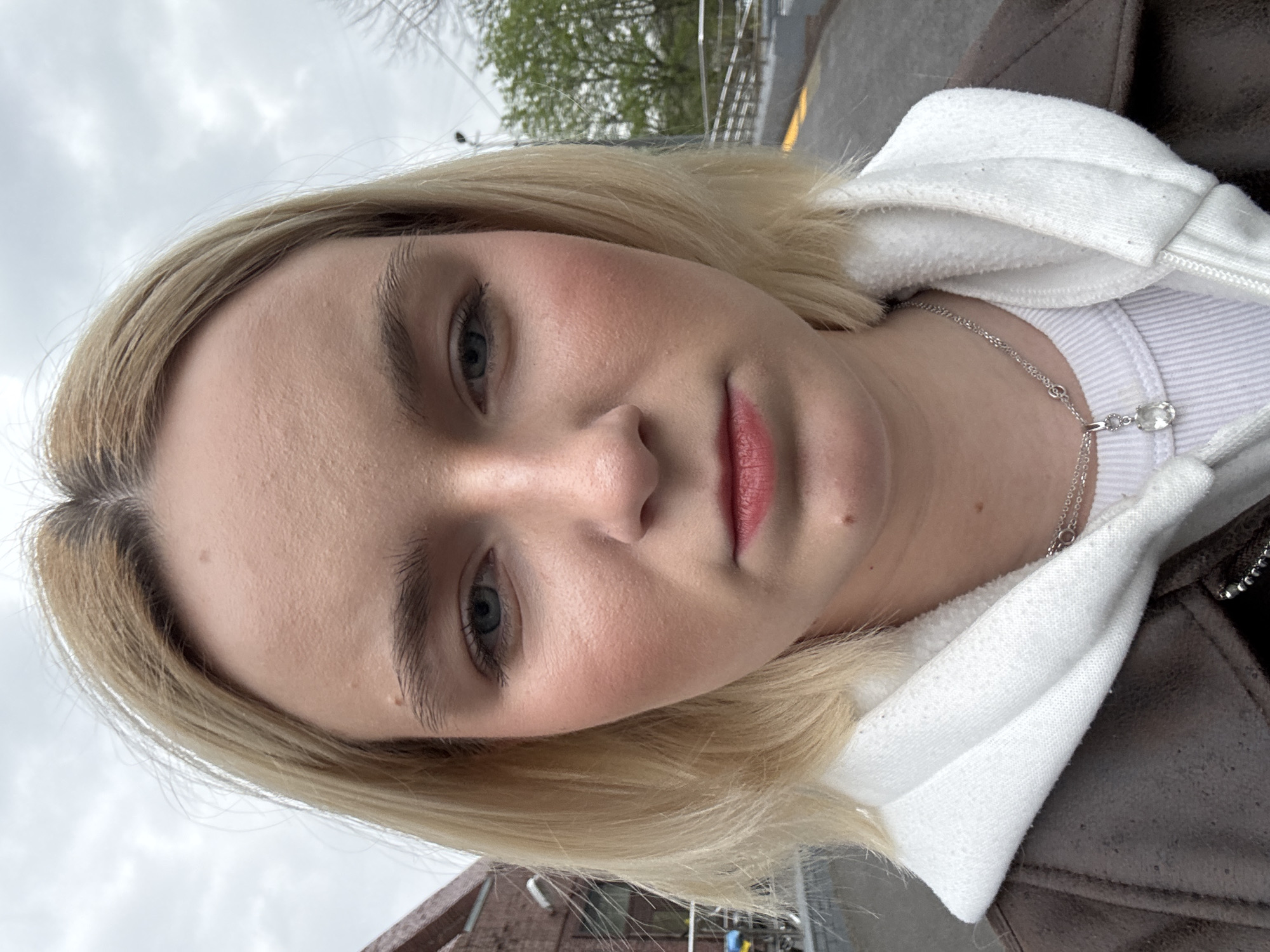 A close-up, slightly angled, photograph depicts a young woman with fair skin, blonde hair, and light blue eyes. She has a white hooded sweatshirt under a brown jacket. A silver necklace with a clear pendant is visible around her neck. The background shows a blurred cityscape with trees and a cloudy sky. A brick building with bicycle parking is visible. The environment suggests an urban setting in a location resembling St. Petersburg, Russia.