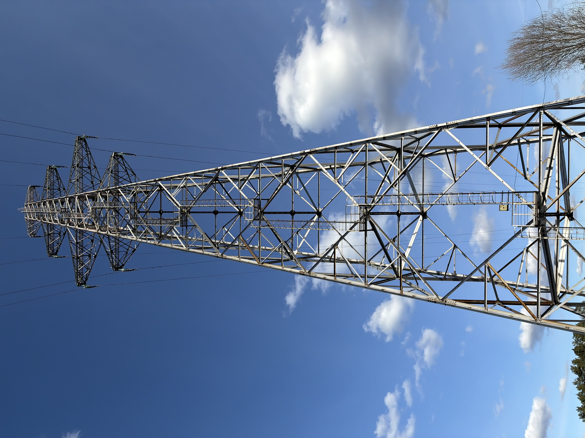 A horizontal, low-angle shot shows an electrical transmission tower against a sky with scattered white clouds. The tower is made of a lattice of interconnected metal beams, forming a robust structure. Electrical wires are strung between the tower and distant towers, which appear smaller in the background. The sky transitions from a darker blue near the top to a lighter blue closer to the bottom of the frame. Vegetation is visible on the lower right corner. The location is the city of Chaykovsky, Russia.