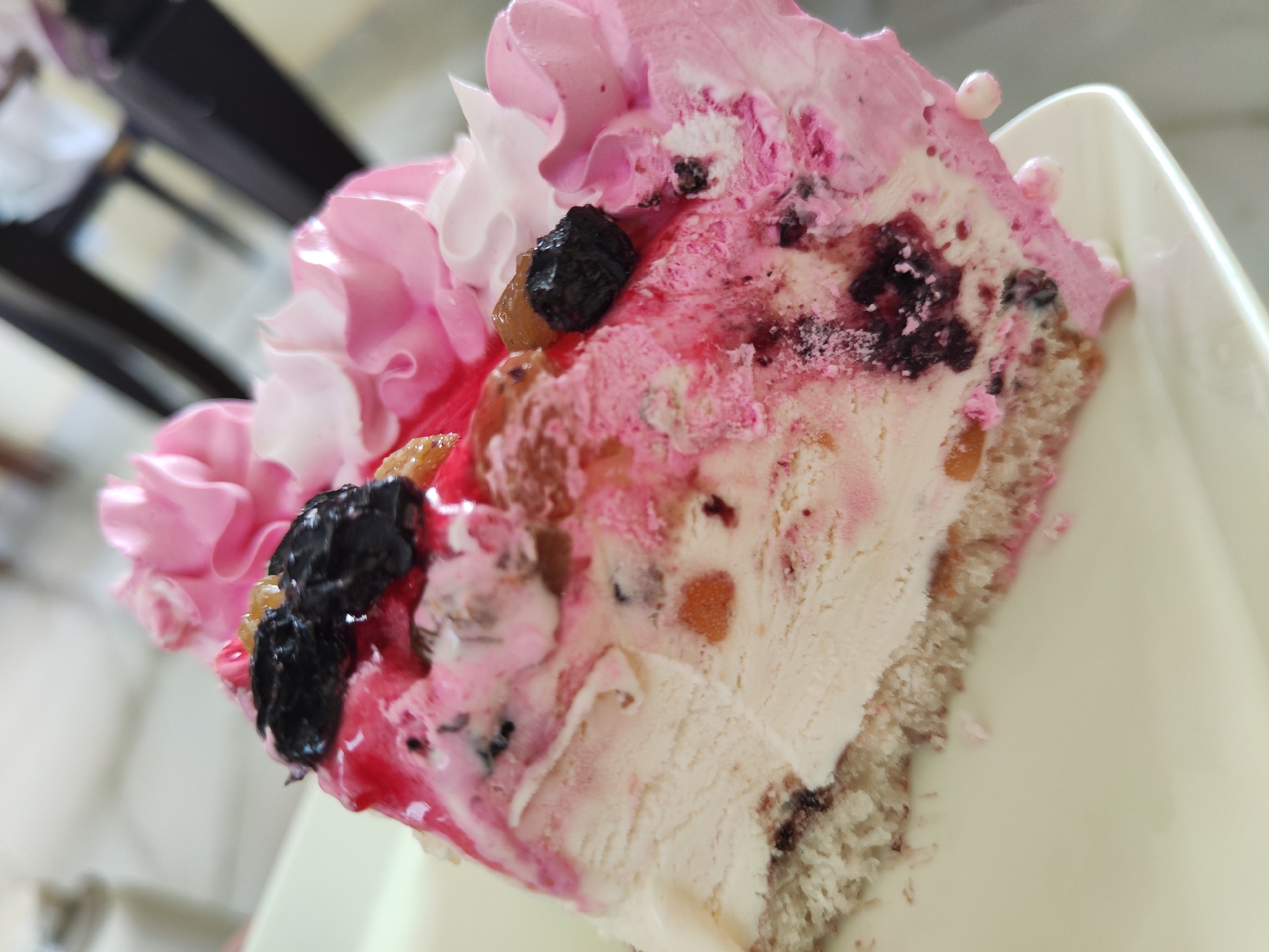 The image shows a slice of cake, likely served on a white plate. The cake features a base of sponge cake, topped with a layer of ice cream, and adorned with pink frosting, white swirls, and scattered blueberries. The scene lacks identifiable markers to specify the city or location.