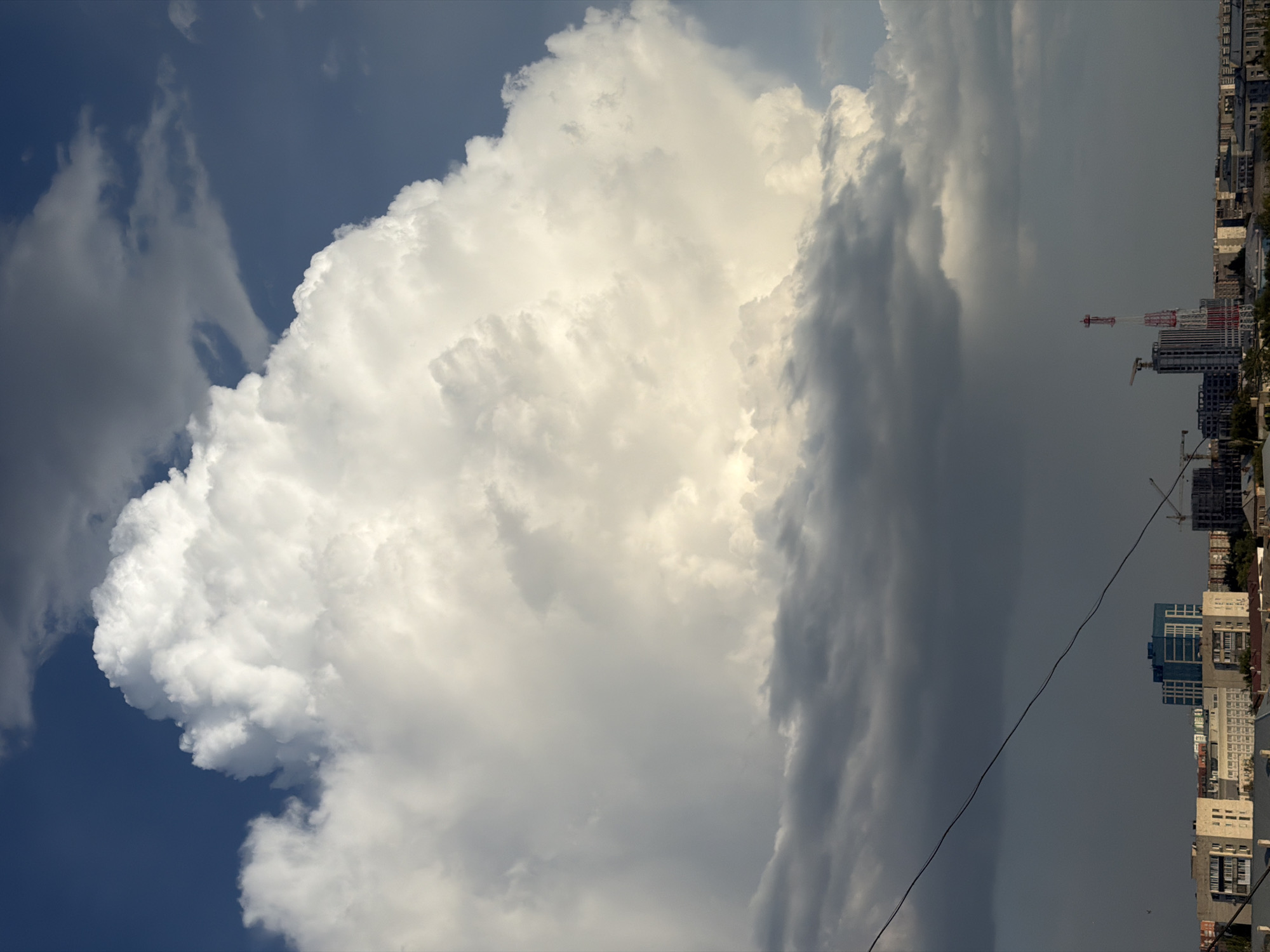 The image depicts a dramatic sky over the city of Pavlodar, Kazakhstan. A massive, bright white cumulus cloud dominates the upper half of the frame, with darker, more ominous clouds forming a band below it. The sun is clearly behind the large cloud, illuminating its edges and creating a luminous effect. Below the clouds, a city skyline is visible, characterized by a variety of buildings, including apartment blocks, office buildings, and what appears to be a construction site with cranes. A prominent red and white communication tower stands out in the mid-ground. A single power line stretches diagonally across the lower portion of the image, connecting to unseen points outside the frame. The overall impression is one of an impending storm, with the stark contrast between the bright cloud and the darkened cityscape emphasizing the atmospheric conditions. There are no people or animals visible in the frame.