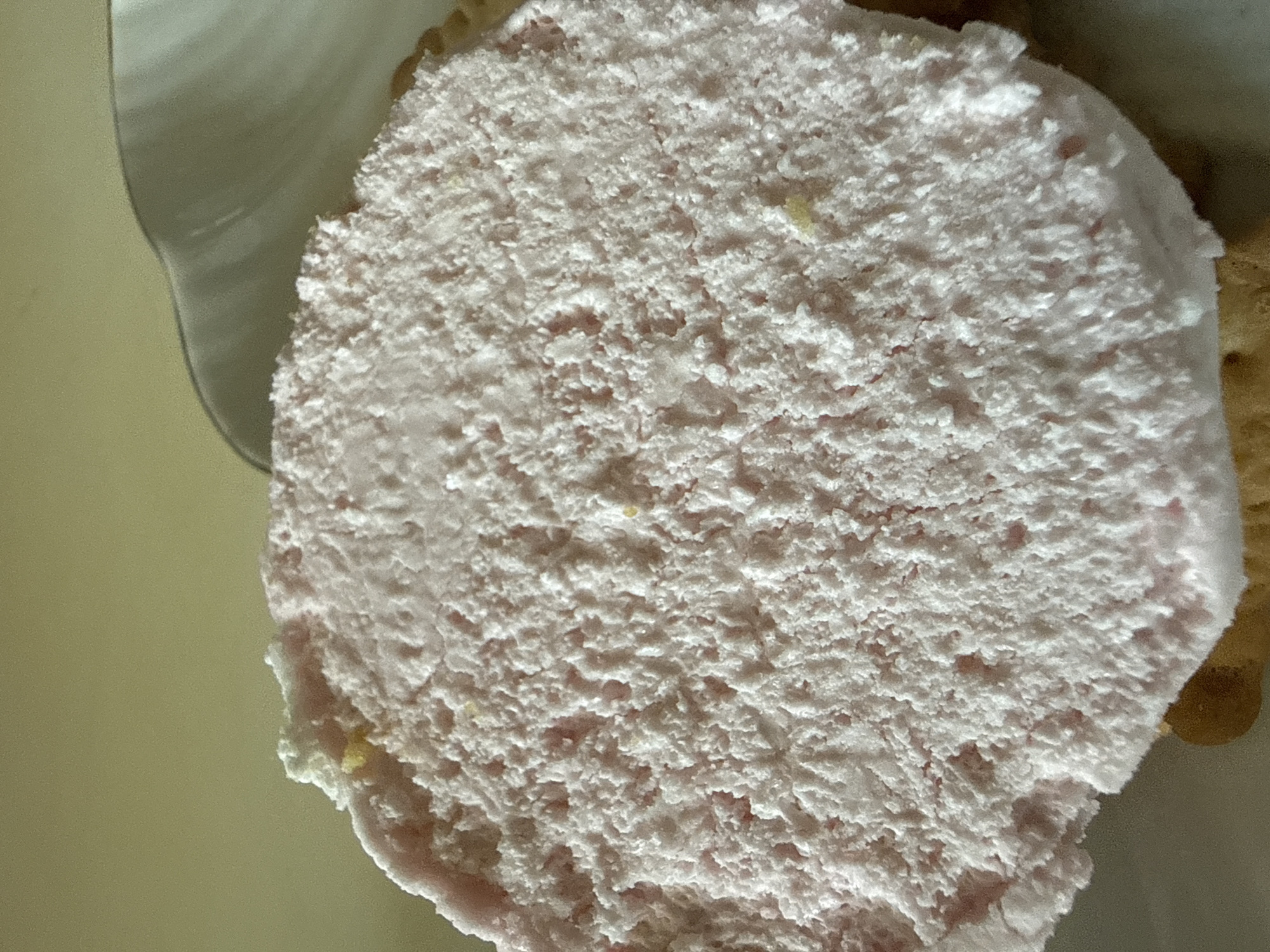 A close-up, overhead shot shows a light pink, fluffy confection on top of a cracker. The confection has a rough, uneven surface with small air bubbles and some lighter white specks. A few small yellow specks are visible within the pink confection. The cracker beneath is a light brown color. A white, curved dish is visible in the upper right corner of the image. The background is a plain, light tan color. The scene is indoors, with no indication of a specific city or location.