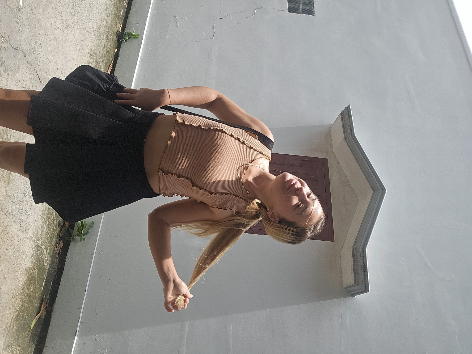 A young woman with blonde hair tied in a ponytail is posing outdoors in front of a white textured wall with a dark brown wooden door and window. She is wearing a peach-colored ribbed crop top and a black pleated skirt. She is holding a lock of her hair in her right hand and her head is tilted back with her eyes closed, as if basking in the sun. The ground is paved and there are a few small plants growing along the base of the wall. The scene suggests a sunny day in Mũi Né, Vietnam, a coastal resort town known for its beaches and sand dunes.
